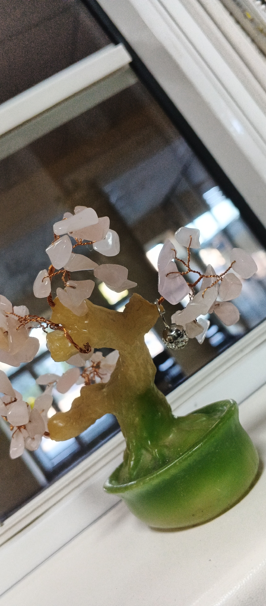 A close-up, angled shot shows a small decorative bonsai tree on a windowsill. The tree has a light brown and green trunk with a green, dish-like base. Its branches are adorned with numerous light pink, irregular-shaped stone "leaves," some appearing translucent. A small, silver-colored charm hangs from one of the branches. The background is out of focus, showing a reflection of windows and the outdoors, with a hint of a dark, textured surface. The scene appears to be indoors, near a window, with the light suggesting it might be daytime. The location is identified as Smila, Ukraine.