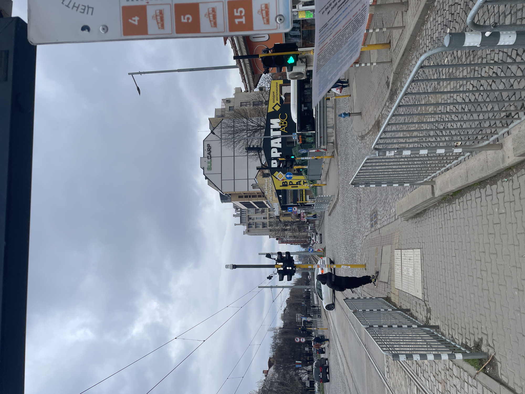 The image depicts a street scene in Sofia, Bulgaria, under a cloudy sky. On the right side of the image, a section of a metal railing is visible, bordering a paved sidewalk with a few rectangular utility covers. A person in a black outfit and white sneakers is walking along the sidewalk. A white car is parked near the curb, alongside tram tracks. Overhead are electrical wires and traffic lights, one displaying a red signal. In the background, buildings with various commercial signage can be seen, including the prominent words "BULGARP" and "РАТИ ABC." Other signage is present in Bulgarian. A tram stop sign with route numbers is visible in the upper left corner. The street is lined with cars and trees.