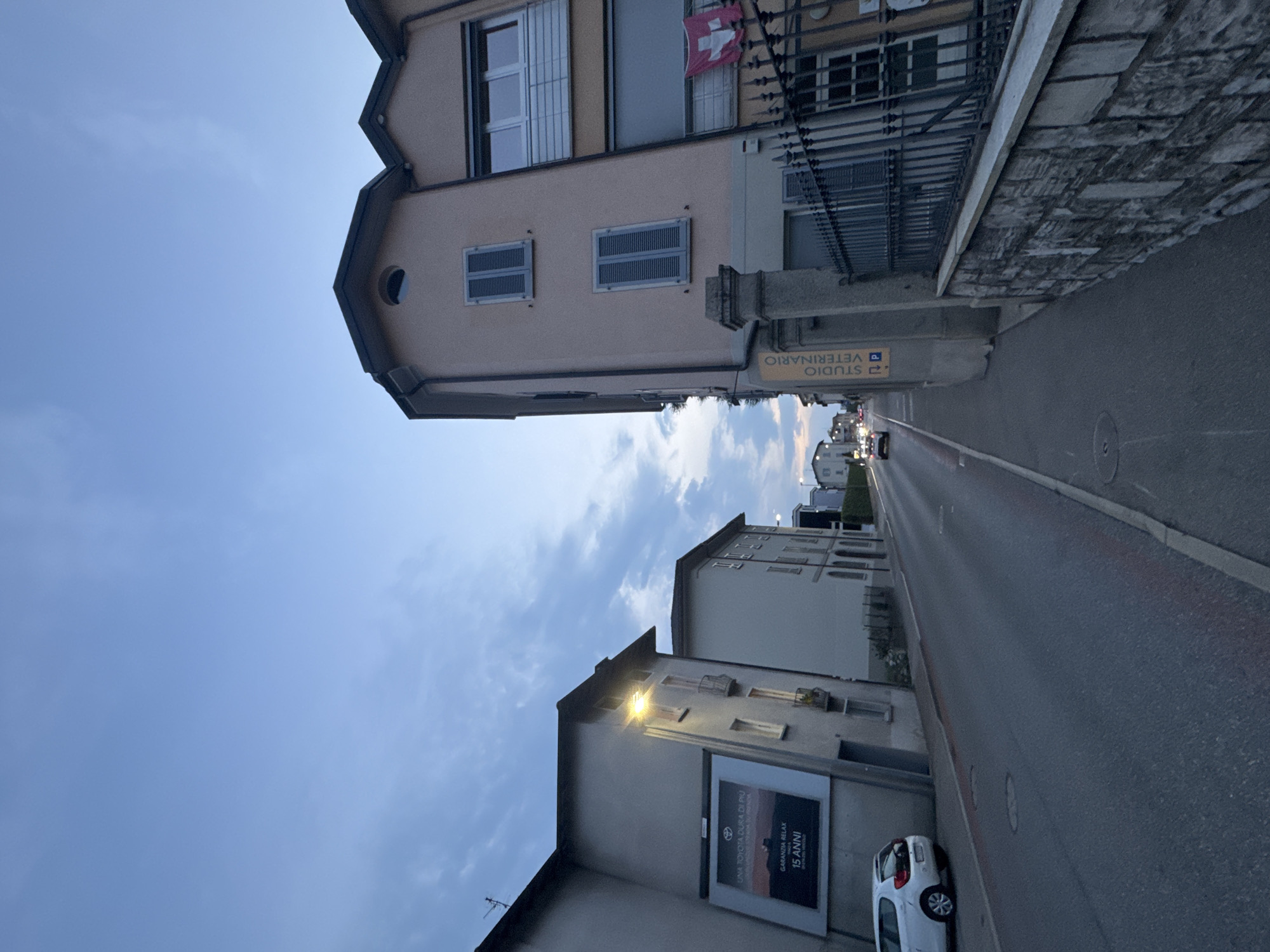 The image depicts a street scene in the city of Balerna, Switzerland, during twilight. On the right side of the street, a light pink building stands with dark shutters on its windows. A Swiss flag is visible hanging from the building. A black metal fence with a stone wall in front of it borders the property. A vertical sign reads "STUDIO VETERINARIO".
On the left side of the street, a grey building features a large billboard advertising "GARANZIA RELAX 15 ANNI". A white car is parked on the left side of the road. The street is paved and appears to be a two-lane road with traffic lights visible in the distance. The sky above is a clear blue with some wispy clouds, suggesting it is either early morning or late evening. The overall atmosphere is quiet and suburban.