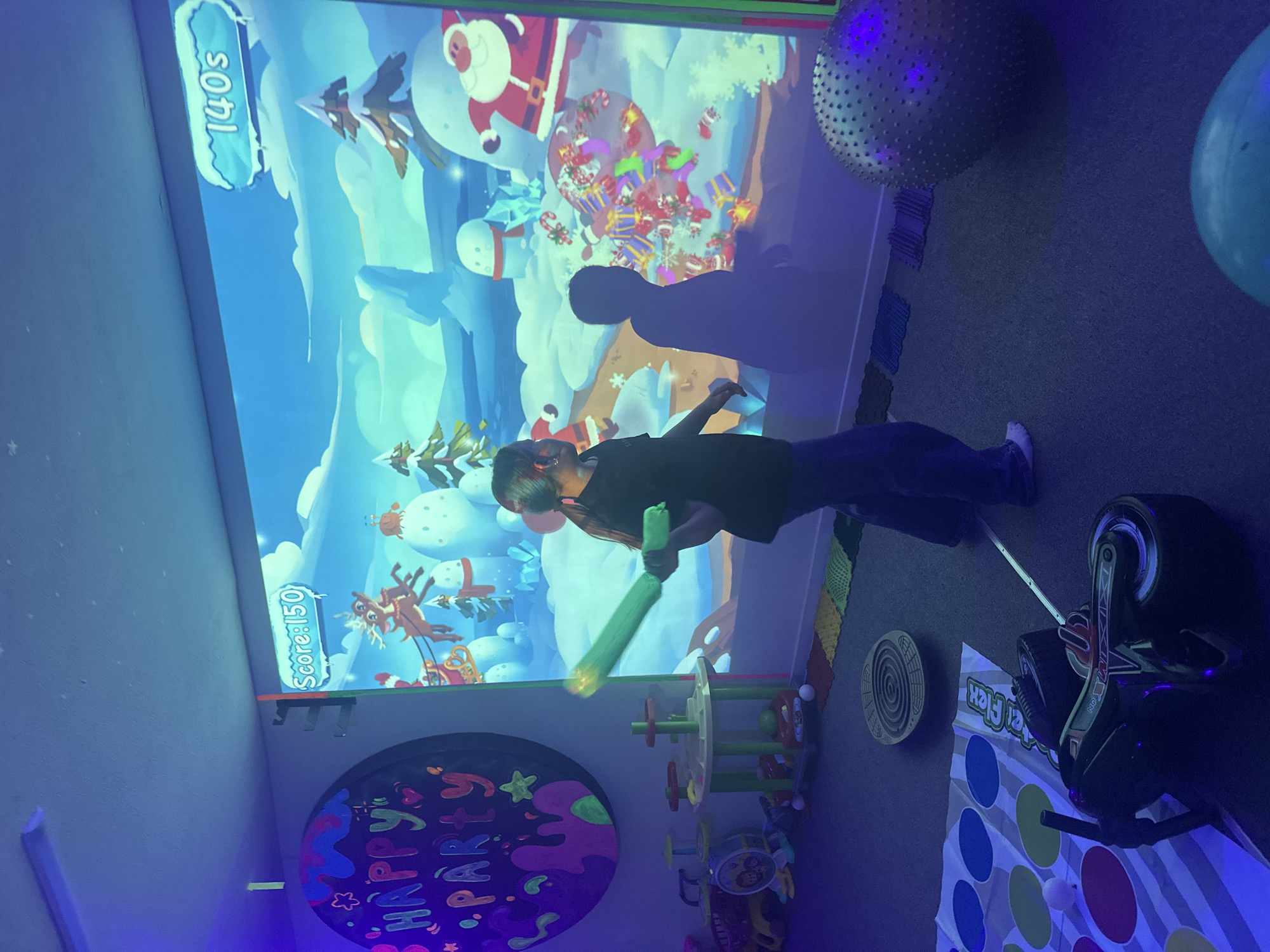 The image depicts a young girl interacting with a projected game in a room illuminated by blacklights. The girl, positioned in the center of the frame, is facing a large projection on the wall, which displays a winter-themed scene with Santa Claus and reindeer. She is holding a green bat-like object, suggesting she is actively participating in the game. The projected game interface shows "Score: 150" and "140s".
The room itself appears to be a recreational or therapy space, evidenced by the various objects scattered on the floor. These include a Twister mat, a balance board, colorful textured mats, a large spiky exercise ball, and a toy motorcycle. A circular decoration with the words "HAPPY PARTY" is visible on the left wall, also highlighted by the blacklights. The lighting is predominantly purple and blue, casting shadows and giving the scene a vibrant, otherworldly glow. The overall environment is child-friendly and designed for interactive play.