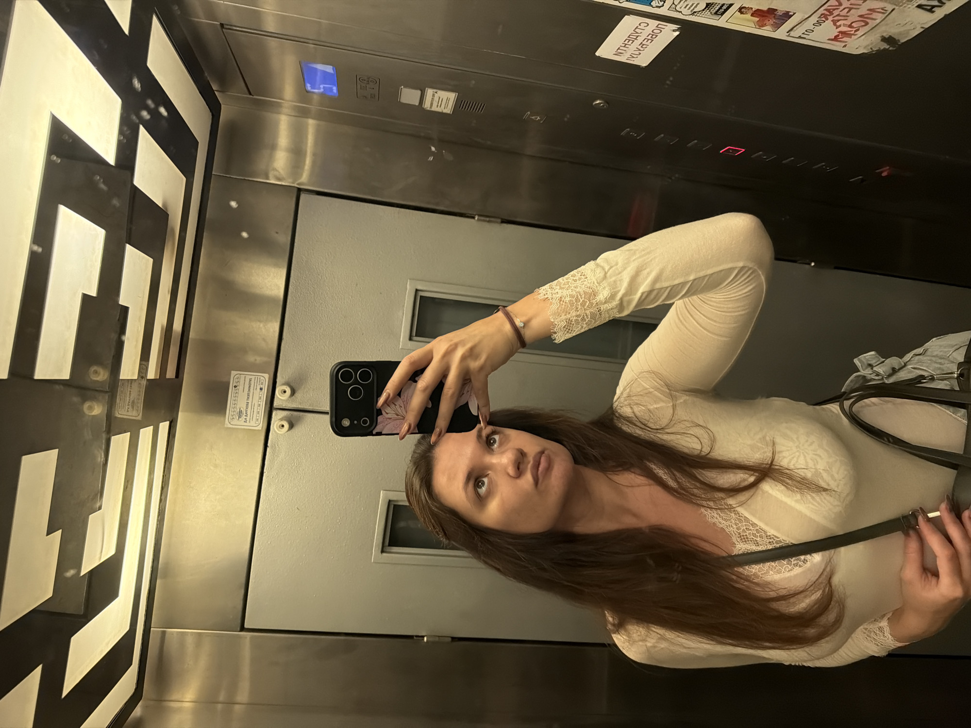 A young woman is taking a selfie in an elevator. She is holding her phone in her left hand, and her right hand is holding the strap of a bag. The elevator has a black and white geometric pattern on the side and metal doors with various labels in Cyrillic script. The elevator control panel with buttons and a display is also visible. There are various stickers above the doors. Based on the Cyrillic writing and general aesthetic, this image was taken in the city of Belgrade, Serbia.