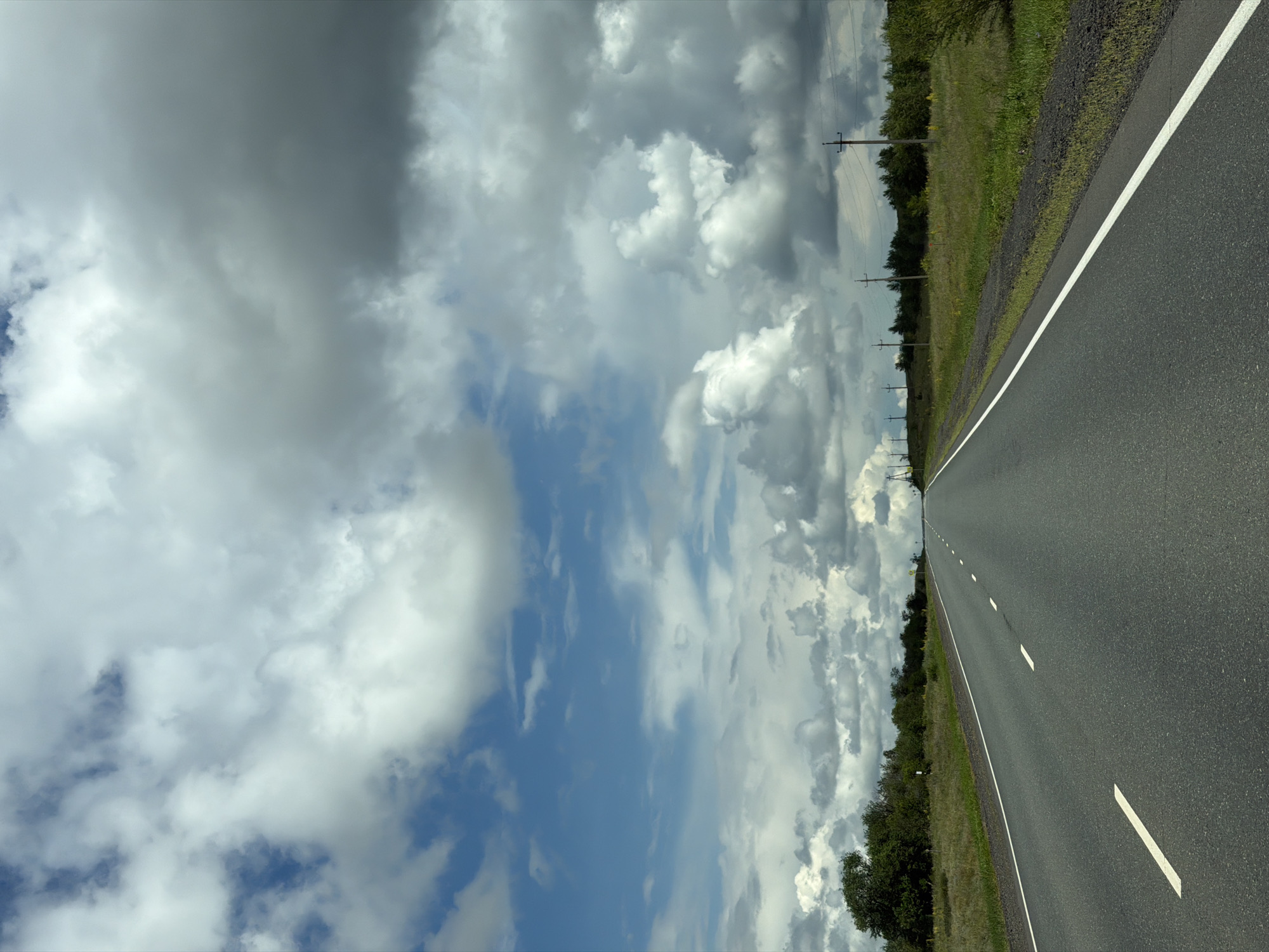 The image depicts an asphalt road stretching into the distance, flanked by grassy verges and a line of trees. The sky is filled with dramatic, dark clouds interspersed with patches of blue sky. A few telephone poles are visible along the roadside. There are no people or vehicles in the frame. The scene suggests a rural or highway setting. The location is the city of Ostrovnoye, Russia.