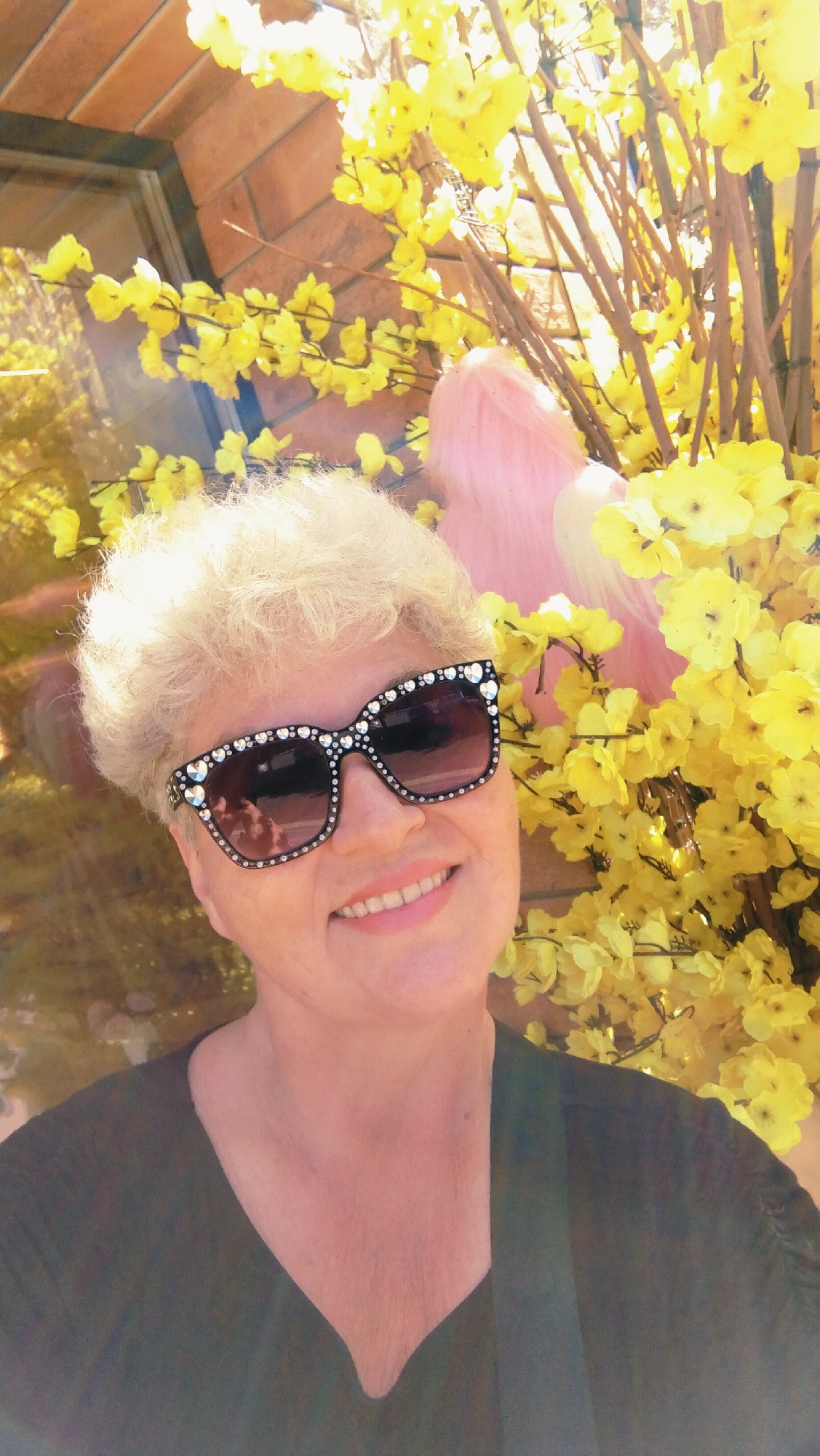 The image shows a close-up, selfie-style portrait of a woman with short, blonde, curly hair and fair skin. She is wearing large, black sunglasses adorned with rhinestones. Her expression is a smile, revealing her teeth. She is wearing a dark, V-neck top. Behind her, there is a display of bright yellow artificial flowers and branches. To the left, a reflection in a window or glass surface shows some of the surrounding environment, including what appears to be a brick wall. A blurry, pink object, possibly a decorative item or a stuffed toy, is visible in the background among the flowers. The overall lighting suggests an outdoor or well-lit indoor setting. The scene appears to be in the city of Horishni Plavni, Ukraine, as implied by the context of the request, though no specific landmarks directly identify this location within the image itself.
