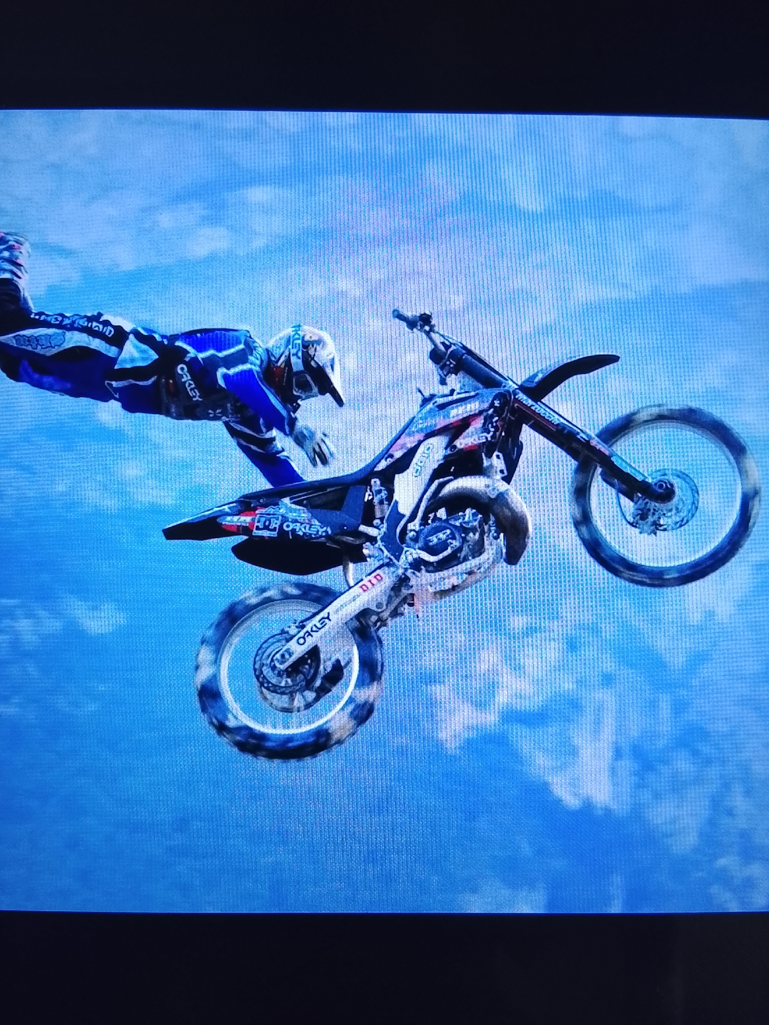 The image shows a motocross rider performing a jump against a blue sky with clouds. The rider is wearing a blue and white racing suit and helmet. The motorcycle is in mid-air, with the rider's legs extended. The background is a textured blue sky with white clouds. The image appears to be a screenshot or a photograph of a video game or animation, as indicated by the pixellated texture and the unnatural lighting. There is no indication of the image being related to Prisel'e, Russia.