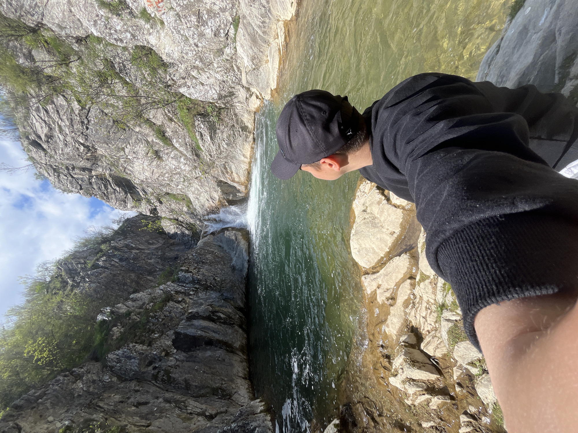 A man stands at the edge of a rocky cliff overlooking a waterfall, likely in the vicinity of Medven, Bulgaria. The man, wearing a black sweatshirt and a black baseball cap, is positioned on the right side of the image and is gazing down at the water below. The waterfall cascades from the top of the image, creating a pool of water surrounded by rocks. On the left side of the frame, there are tall cliffs with sparse vegetation. The sky is visible in the upper left corner, revealing a few clouds.