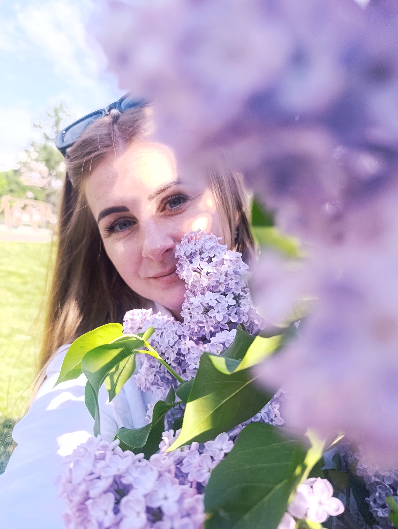 A woman with long brown hair and sunglasses on her head holds a bunch of purple lilacs close to her face. She is wearing a white shirt. The background shows a grassy area and some trees. The location is Unknown, Unknown.