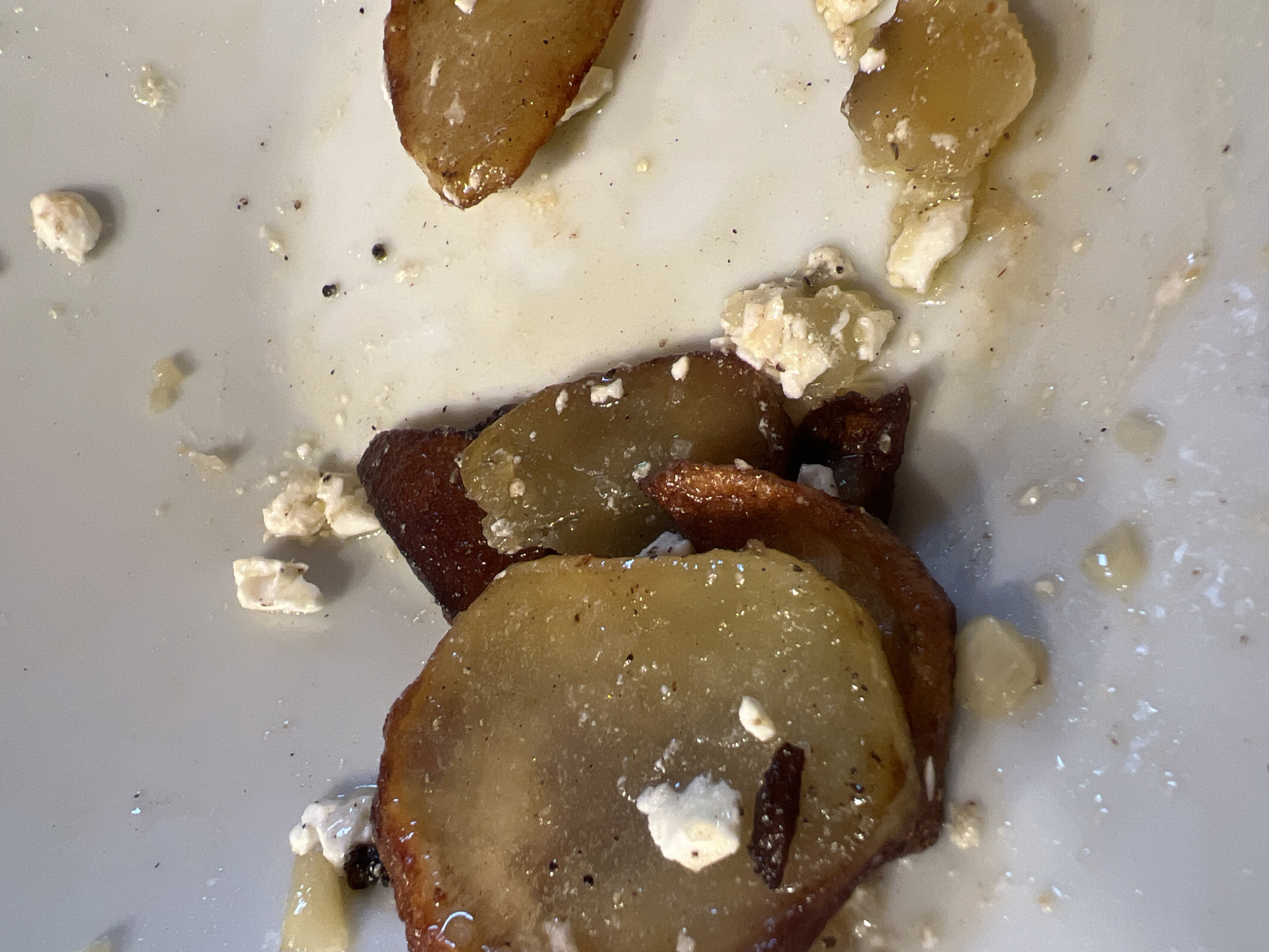 The image shows a close-up of a white plate with leftover food. The food consists of several slices of what appear to be baked or fried fruit, possibly pears or apples, which are golden brown and glistening. Scattered among the fruit slices are small, white crumbles of cheese, likely feta. There are also a few darker, possibly caramelized, pieces of fruit. The plate is slightly soiled with food residue and oil. The lighting is bright, causing some specular highlights on the food and plate. The background is a blurred white surface, indicating the plate is the primary subject. The image appears to be a still life of a partially eaten meal. No people, city, or environmental details are visible.