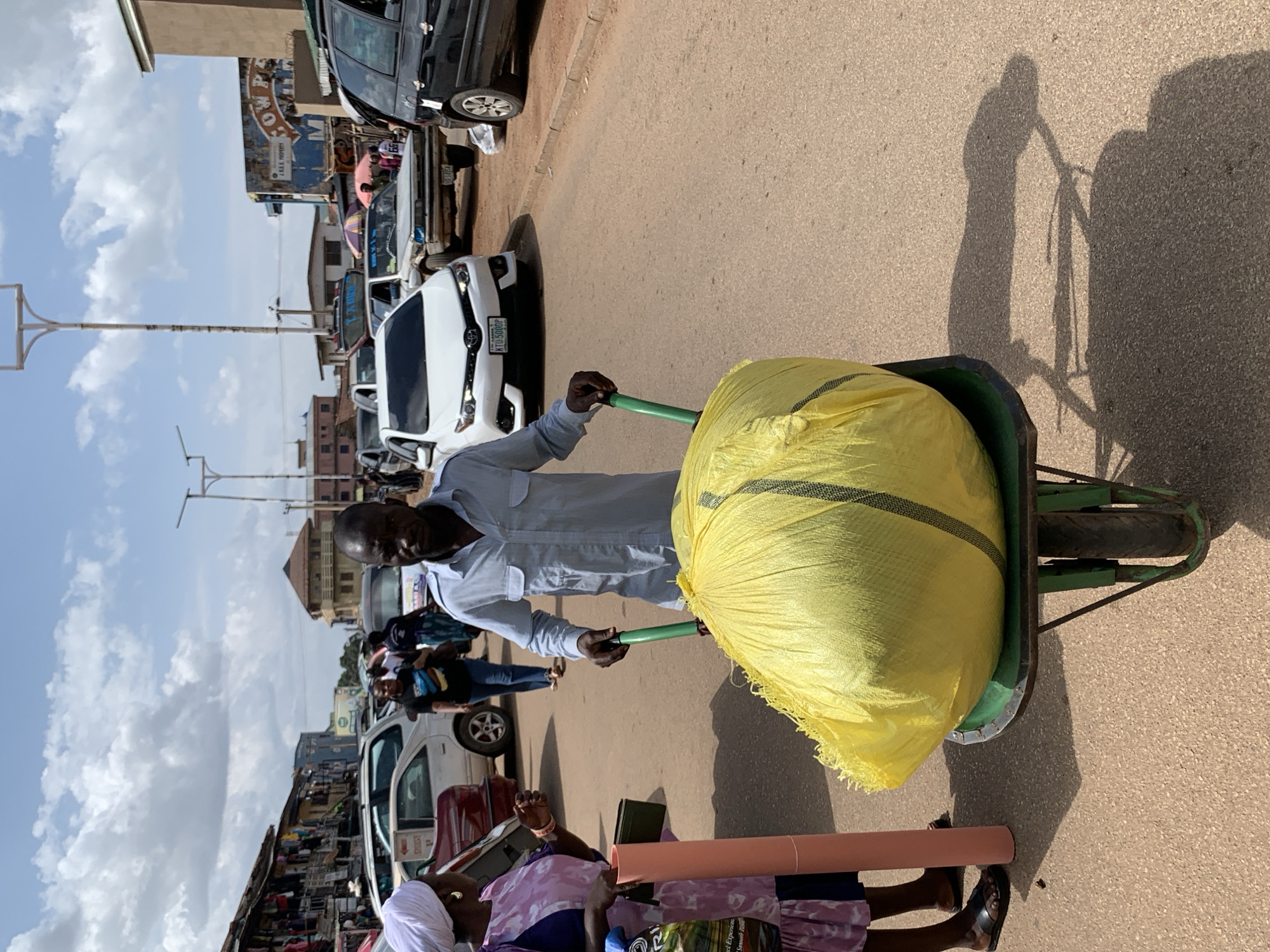 A man in a light blue shirt pushes a wheelbarrow filled with a large, bright yellow bag. The wheelbarrow has green handles and a green frame with two wheels. The scene appears to be a busy street market. In the background, there are several cars, including a white Toyota with a license plate that reads "KTU 5000P". A black SUV and other vehicles are also visible. Storefronts with various goods are lining the street. The sky is partly cloudy, suggesting it is daytime. A woman wearing a pink and purple patterned dress and a white head covering is visible in the foreground, lying on the ground with a long, pink cylindrical object next to her. The overall atmosphere suggests a vibrant and active marketplace. The image is oriented vertically.