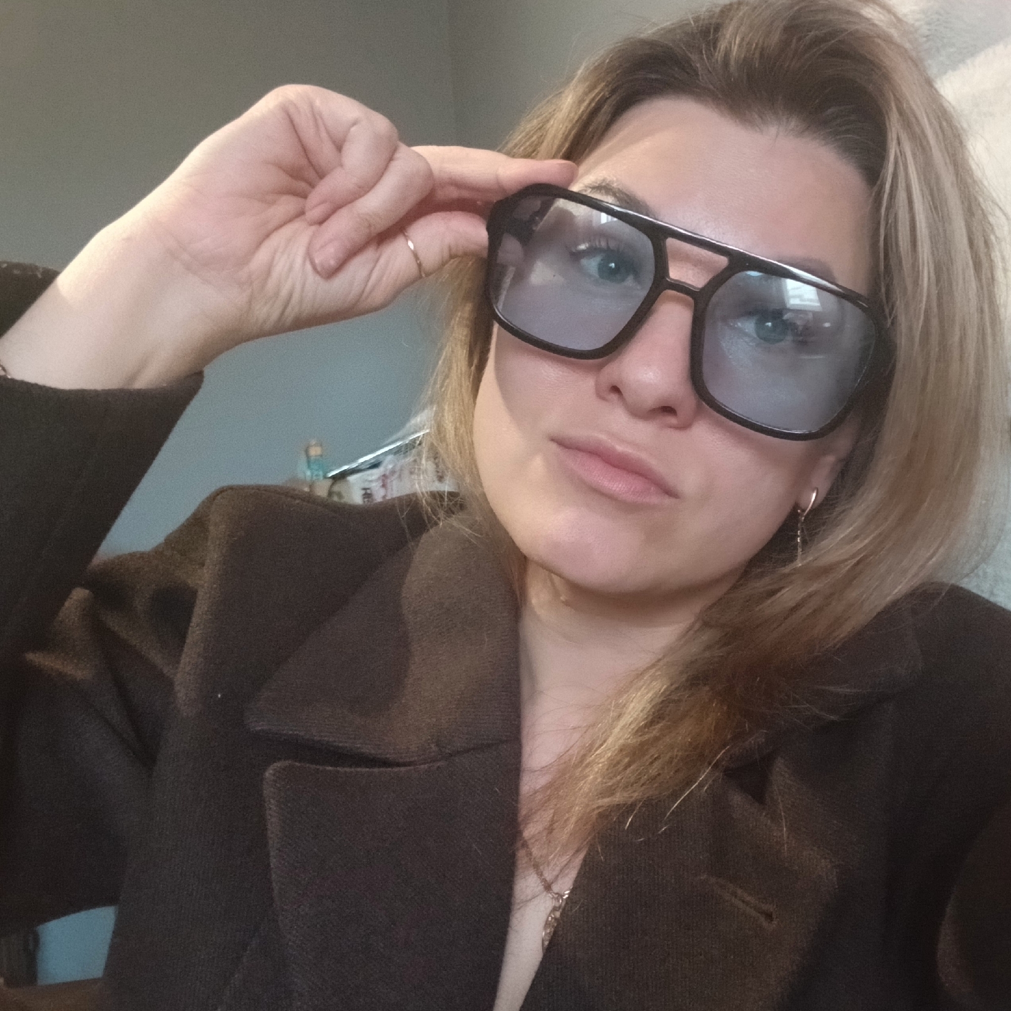 A woman with shoulder-length blonde hair, wearing a dark brown blazer, poses indoors with her right hand touching the upper right corner of her large, black-framed, tinted sunglasses. She is looking at the camera with a serious expression. She wears a small ring on her right ring finger and a dangling earring on her left ear. A necklace is visible at her neckline. The background is out of focus and shows a light blue wall and some indistinct objects on a shelf or table.
There is no information in the image that would indicate the location is in the city of Izhevsk, Russia.