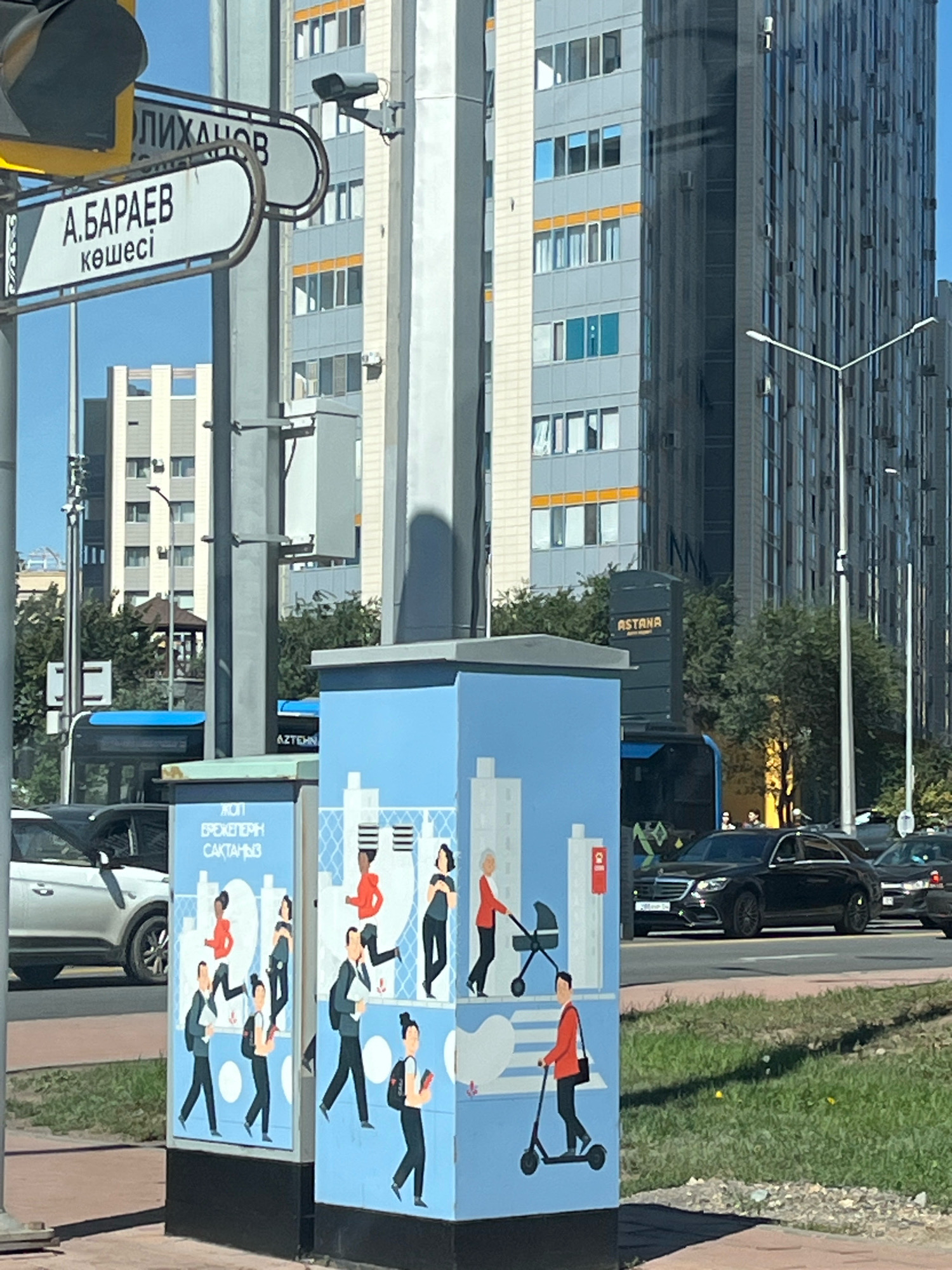 The image shows a street scene in Astana, Kazakhstan, with a focus on a light blue utility box adorned with a mural. The mural depicts stylized figures walking, riding scooters, and pushing strollers against a backdrop of simplified city buildings. The street is paved, with a grassy median strip to the right. Several modern, multi-story buildings with glass facades and balconies dominate the background. A traffic light and street signs are visible on the left, with one sign reading "А. БАРАЕВ көшесі" (A. Baraev Street) and another partially obscured Cyrillic inscription. Several vehicles, including a black Mercedes sedan, are on the road. Trees with green foliage are present between the buildings and the street. The sky is clear and blue.