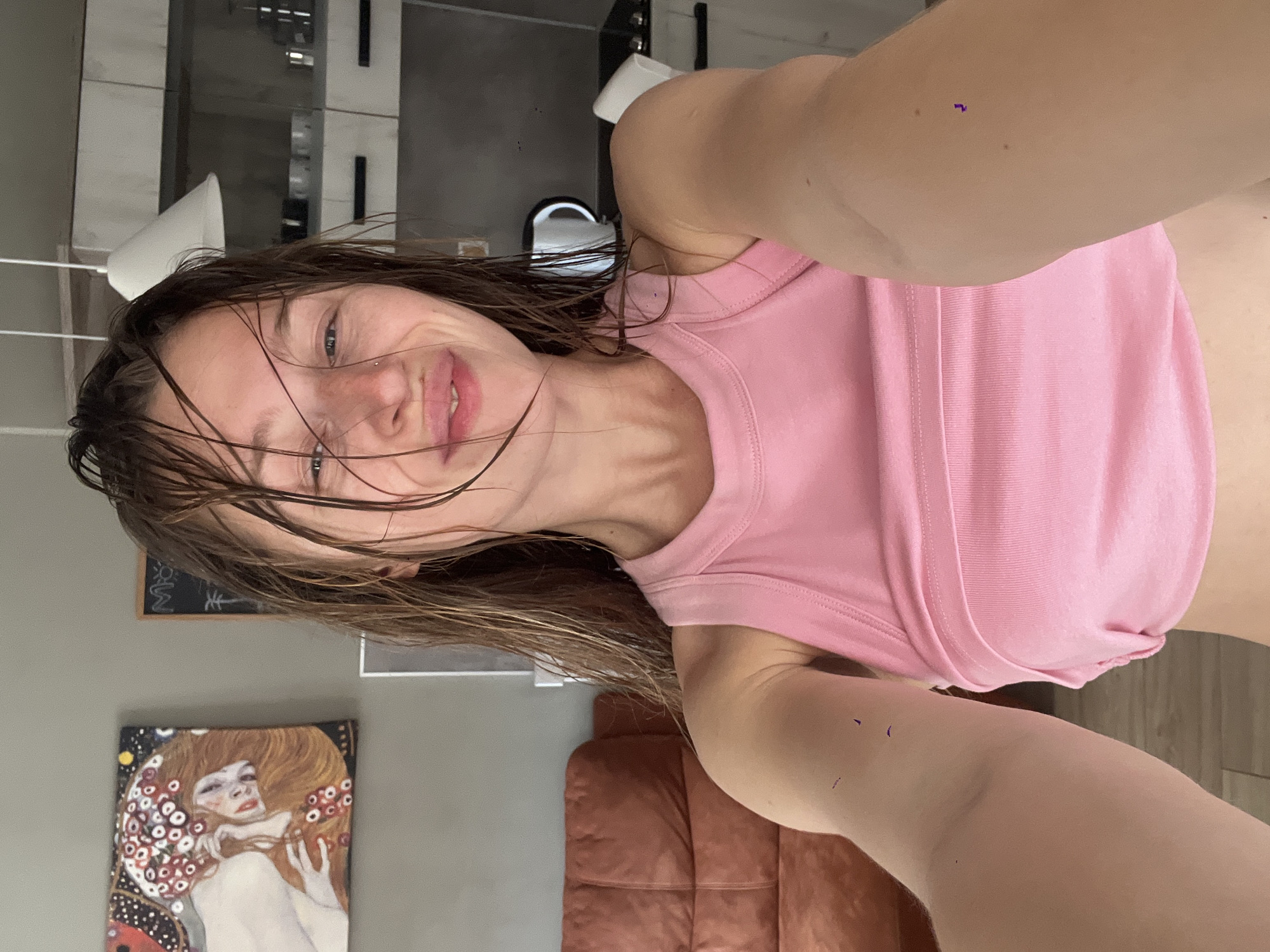 A woman is taking a selfie indoors. She has wet, brown hair and is wearing a pink tank top. Her face is partially obscured by her hair. Behind her, there is a grey wall with a framed artwork depicting a woman. There is also a brown leather couch visible in the background. The location of Rafailovići, Montenegro, cannot be determined from the image.