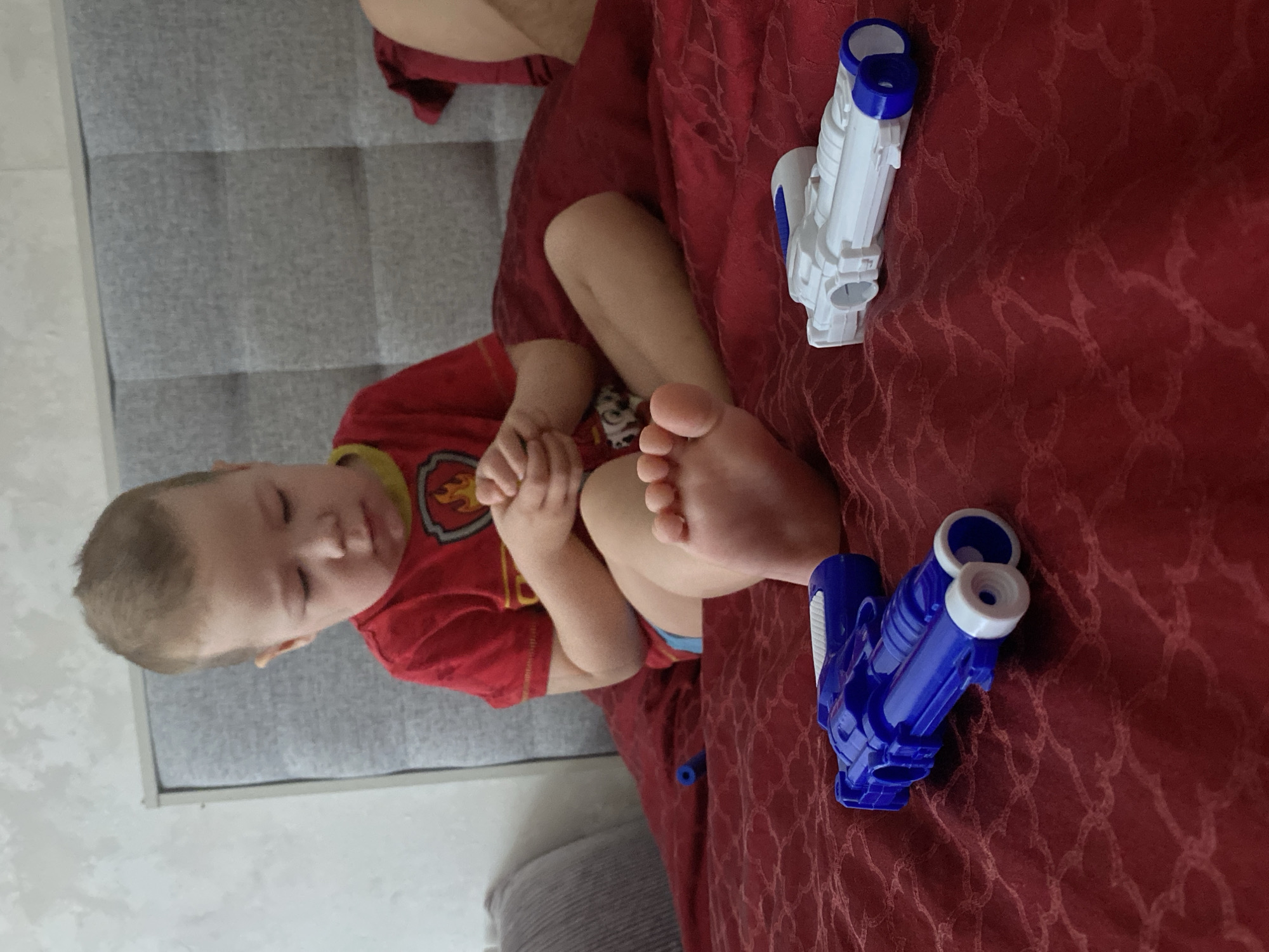 A young child, appearing to be a toddler or preschooler, is captured in a vertical, portrait-oriented image. The child is sitting on a bed or couch, with their knees bent and feet tucked close to their body. Their hands are clasped together in front of them. The child is wearing a red short-sleeved t-shirt with a yellow trim around the neck and sleeves. A graphic, resembling a yellow and orange shield or emblem, is visible on the front of the t-shirt, possibly indicating a character from a children's show. Blue shorts or underwear peek out from beneath the red shirt.
The child's eyes are closed, and their expression is peaceful, suggesting they might be sleeping, resting, or simply closing their eyes. Their light brown hair is short.
The child is situated on a surface covered by a rich, red fabric, likely a bedspread or blanket, which features a subtle, embossed or woven circular pattern throughout. To the child's right, forming a background element, is a grey upholstered headboard or backrest, consisting of three distinct panels.
Two toy blasters or water guns are placed on the red fabric, near the child's feet. One is predominantly white with blue barrels and blue accents, while the other is primarily blue with white barrels and white accents. Both guns appear to have a similar design, featuring double barrels.
The lighting in the scene is soft and ambient, characteristic of an indoor setting. No specific text is visible in the image.