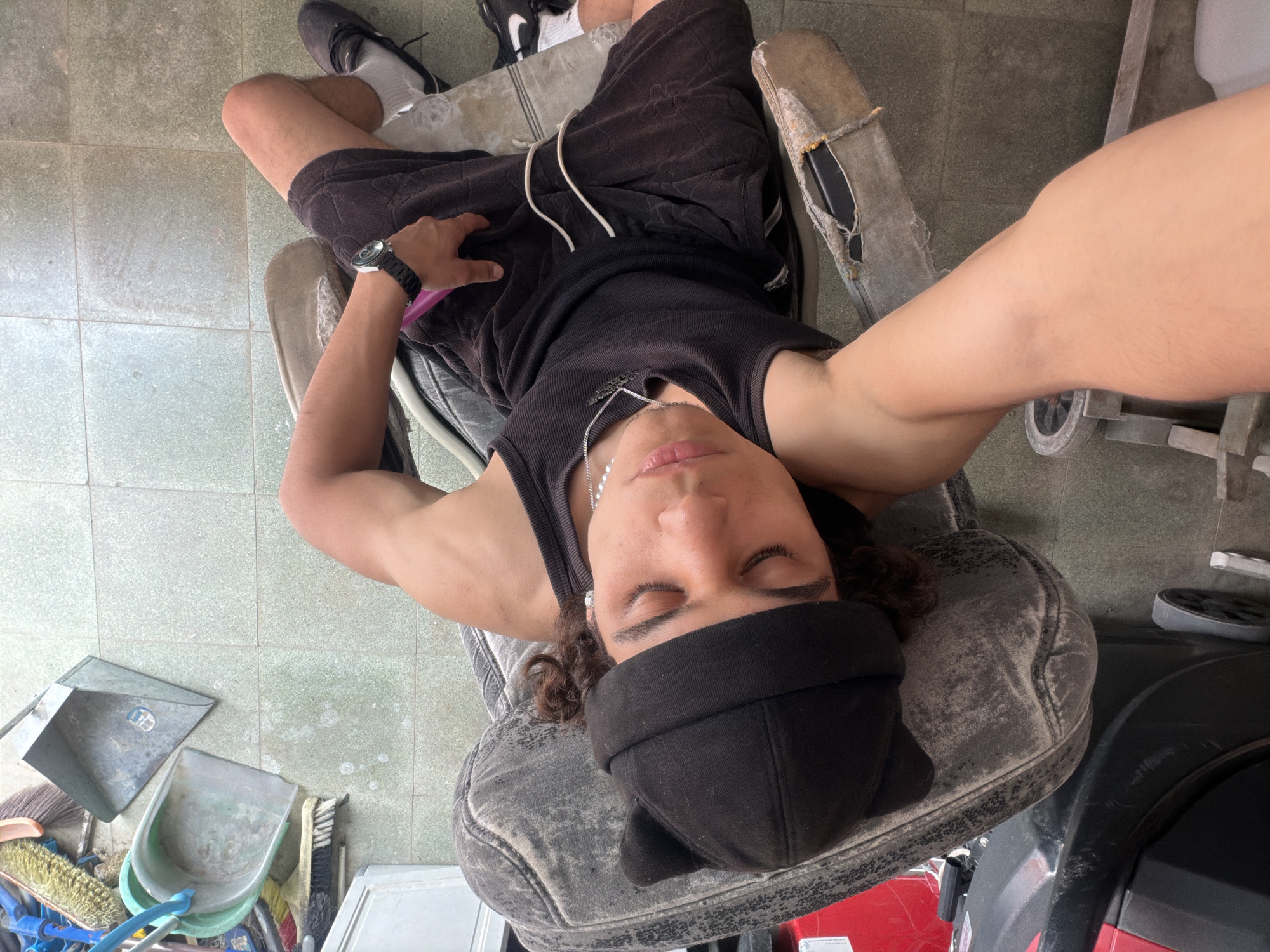An upward-angled full shot captures a person of indeterminate gender, with dark curly hair and tan skin, resting in a worn, light grey or faded black office-style chair. Their eyes are closed, and they wear a black tank top, black shorts with a white drawstring, a black beanie with ear-like protrusions, and a silver chain necklace. A black watch is visible on their left wrist, which also holds a pink or purple object, possibly a phone.
The person's left foot wears a black and white Nike sneaker with white socks, while the right foot sports a well-worn, light grey slip-on shoe with torn fabric. The chair itself shows significant signs of wear, with ripped fabric on the armrests and seat.
The setting appears to be an outdoor or semi-outdoor area, possibly a garage, patio, or utility space in Hof HaCarmel Regional Council, Israel, given the tiled floor that is visibly dirty and stained. To the left of the person, various cleaning supplies are scattered on the floor, including two metal dustpans (one silver, one light green/blue) and several brooms and brushes. To the right, part of a red object, possibly a vehicle or piece of equipment, is visible, along with one of the chair's wheels. The lighting suggests it is daytime. The overall mood is relaxed, with the person seemingly napping or resting.
Visible text includes a partial "NIKE" logo on the left shoe. Other text on a silver dustpan and a white label on the red object is unreadable due to blurriness or distance.