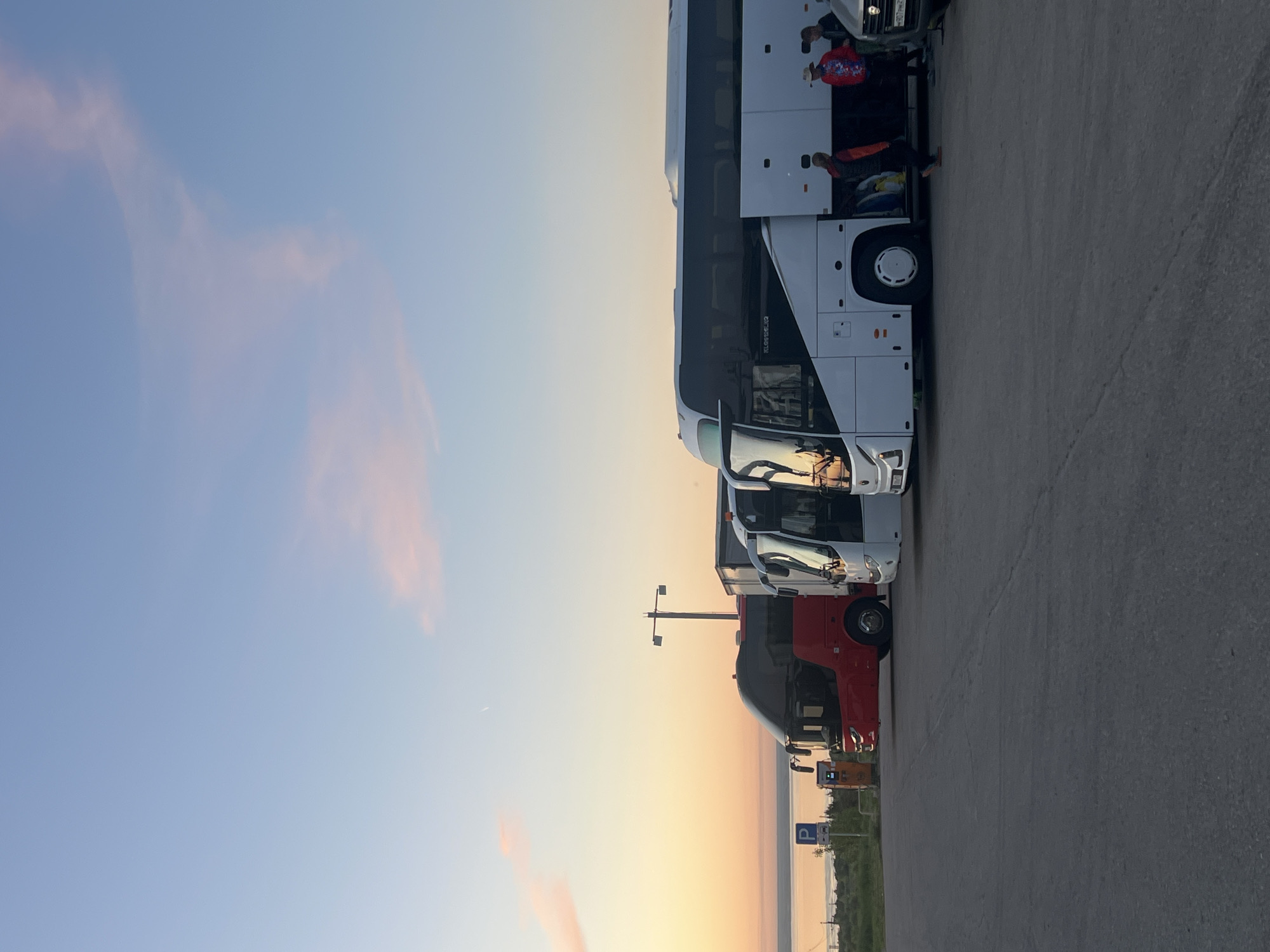 The image shows a row of buses parked at a rest stop or parking area in what seems to be Khvalynsk, Russia. The sky is light, suggesting sunrise or sunset. People are visible near the buses, possibly disembarking or preparing to board. The environment includes a paved area, a few parking signs, some distant trees, and a glimpse of what might be a body of water in the background. The scene captures a transient moment of travel.