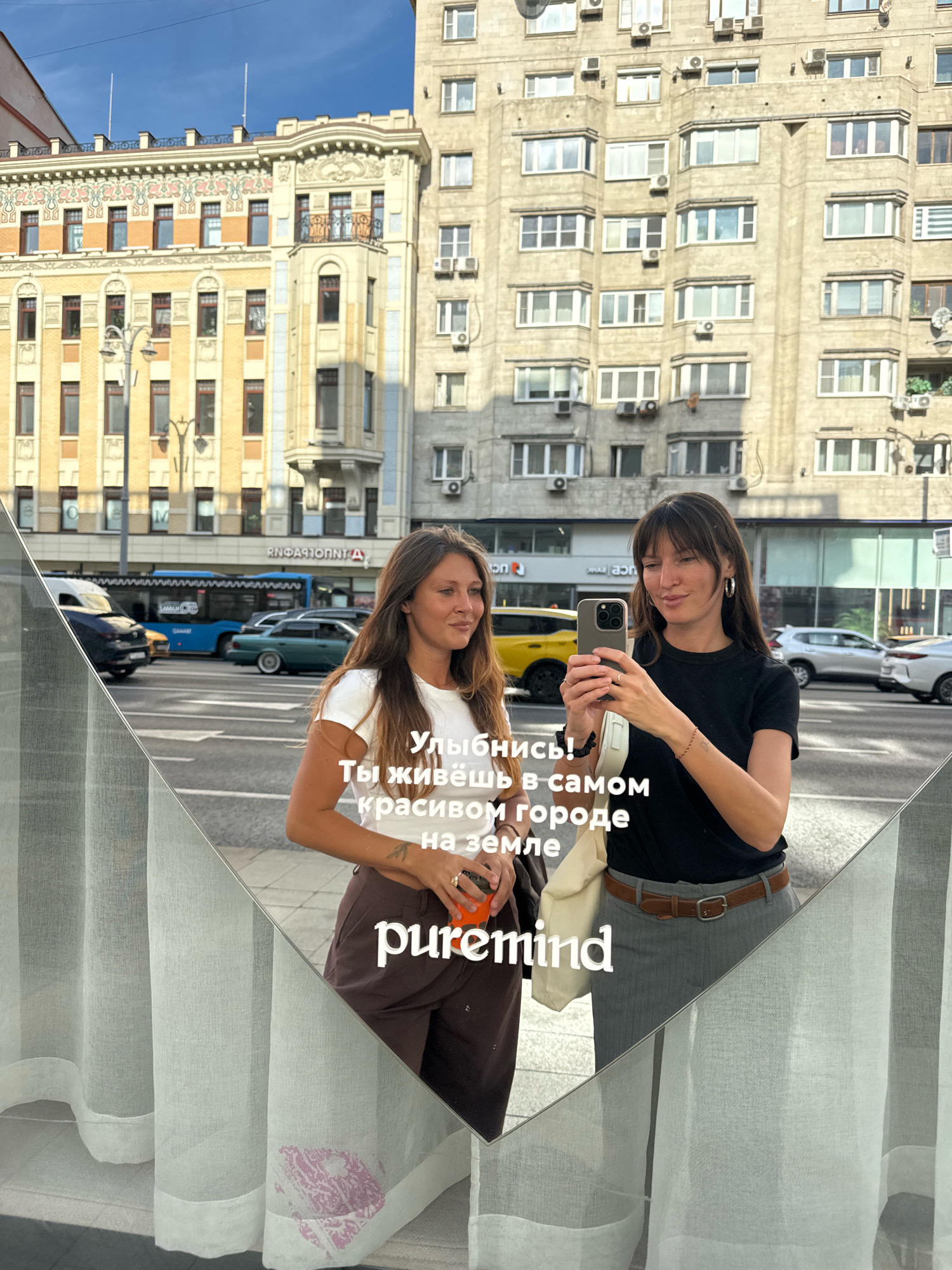 The image shows a reflection in a mirror of two young women on a city street in Moscow, Russia. The woman on the left has long brown hair and is wearing a white t-shirt and brown pants. The woman on the right has dark brown hair and is wearing a black t-shirt and grey pants with a brown belt. She is holding a smartphone and taking a selfie. Text overlays are visible on the mirror in Russian, reading "Smile! You live in the most beautiful city on earth" and "puremind". In the background, there are buildings, cars, and a bus. The architecture suggests an urban environment characteristic of Moscow.