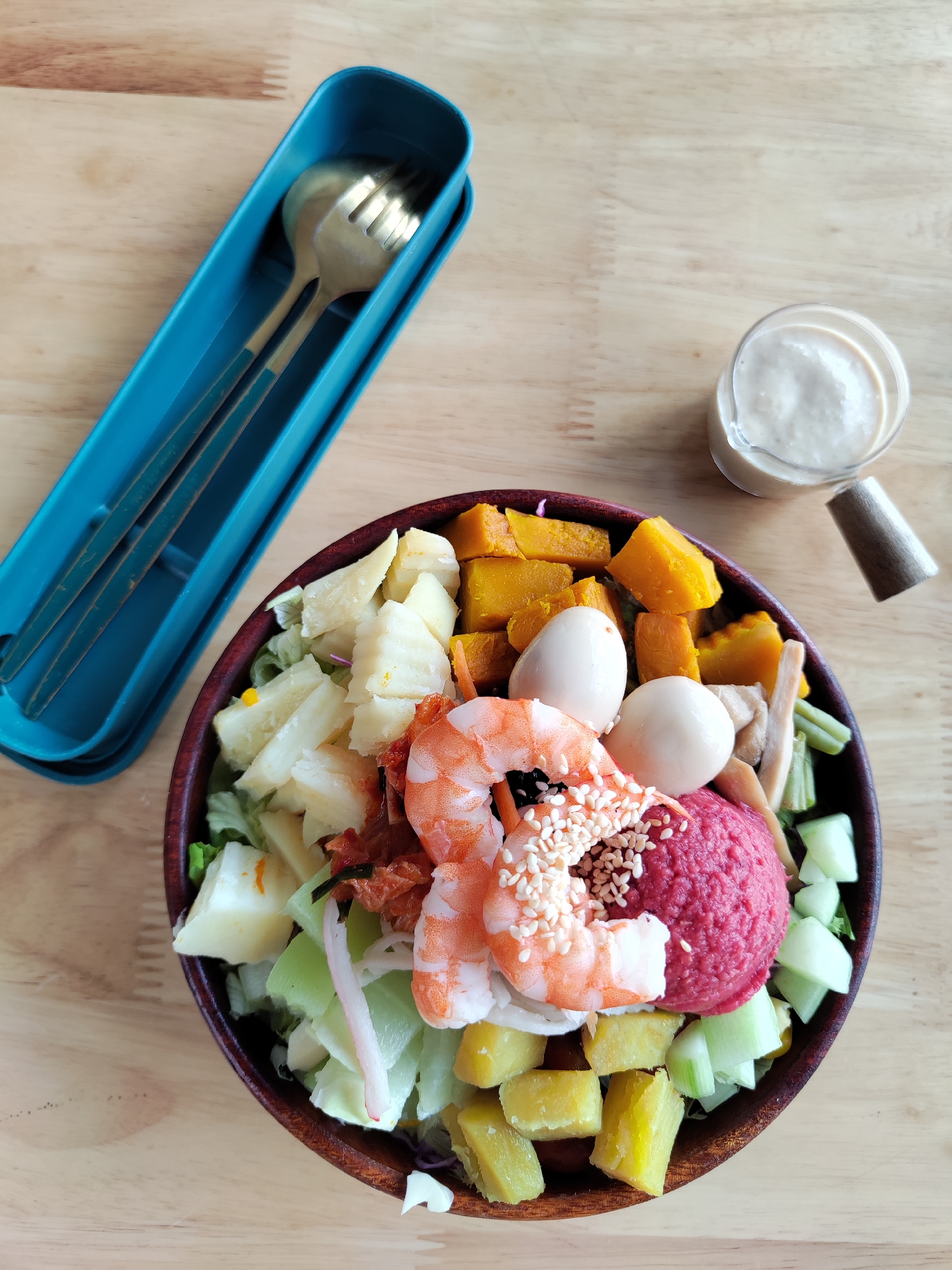 On a light brown wooden table are a bowl of salad, a set of cutlery in a blue case, and a small jar of sauce. The salad bowl contains cooked shrimp topped with sesame seeds, two hard-boiled quail eggs, a scoop of beetroot hummus, chunks of pumpkin, cubed sweet potatoes, and diced white vegetables, possibly jicama or potatoes, all arranged on a bed of mixed greens. The cutlery set in the blue case consists of a spoon and a fork. The small jar contains a creamy white sauce with a wooden handle attached to the side. The photo was likely taken in Da Lat, Vietnam, as the salad ingredients are commonly found in the region's cuisine. The scene suggests a meal setting, ready to be consumed.