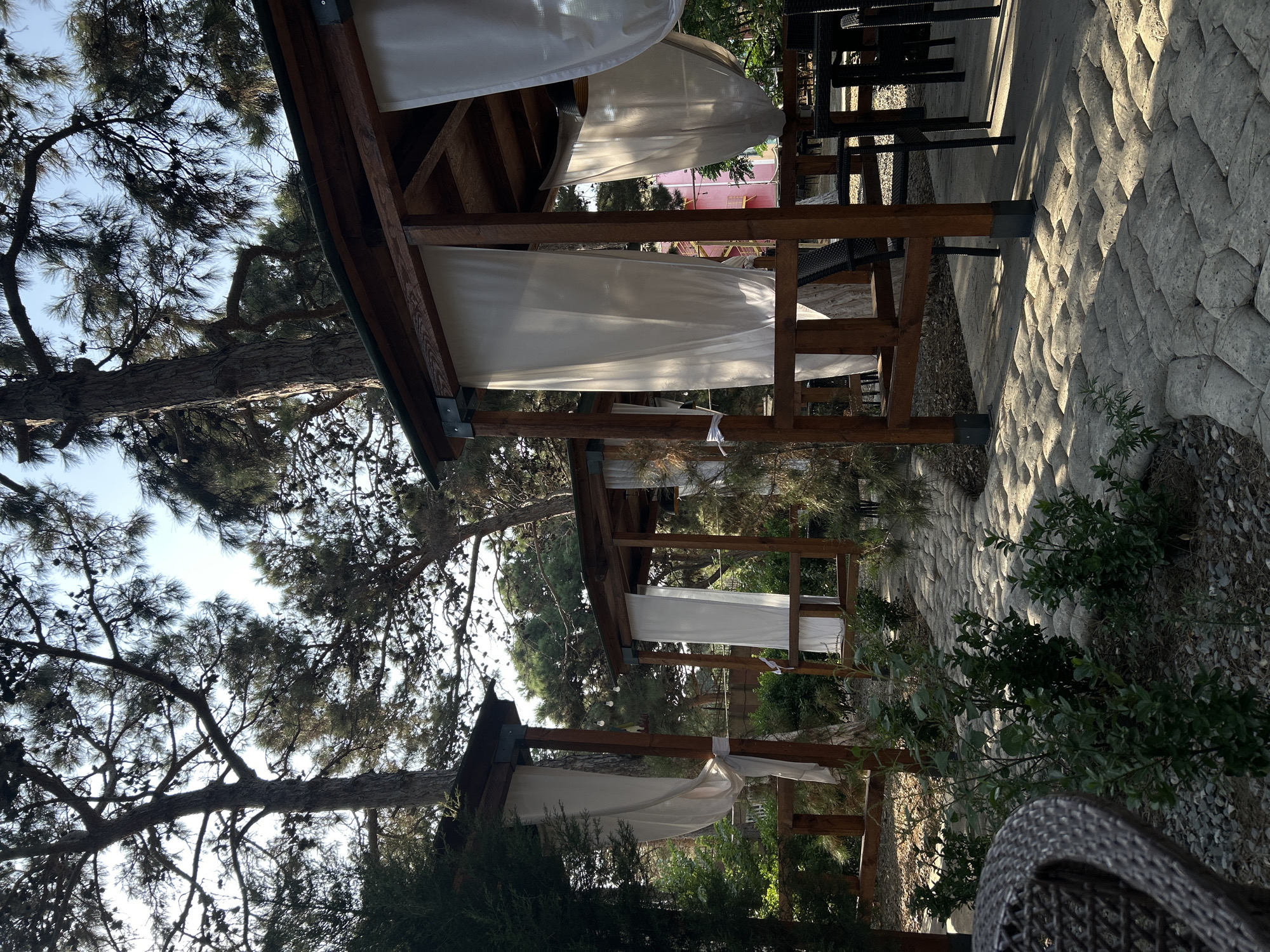 A tranquil outdoor seating area is depicted, likely a cafe or restaurant, set amidst lush greenery and tall trees. The scene features several wooden gazebos with white drapes, suggesting an open-air dining experience. The ground is paved with irregular stone tiles, creating a naturalistic pathway. In the foreground, a portion of a woven chair is visible. Beyond the gazebos, tables and chairs are arranged, hinting at a place for relaxation and dining. The lighting suggests it's daytime, with sunlight filtering through the trees, casting dappled shadows. The architecture and surrounding vegetation evoke a sense of calm and connection with nature. The specific location in Baku, Azerbaijan, cannot be determined solely from the visual information provided.