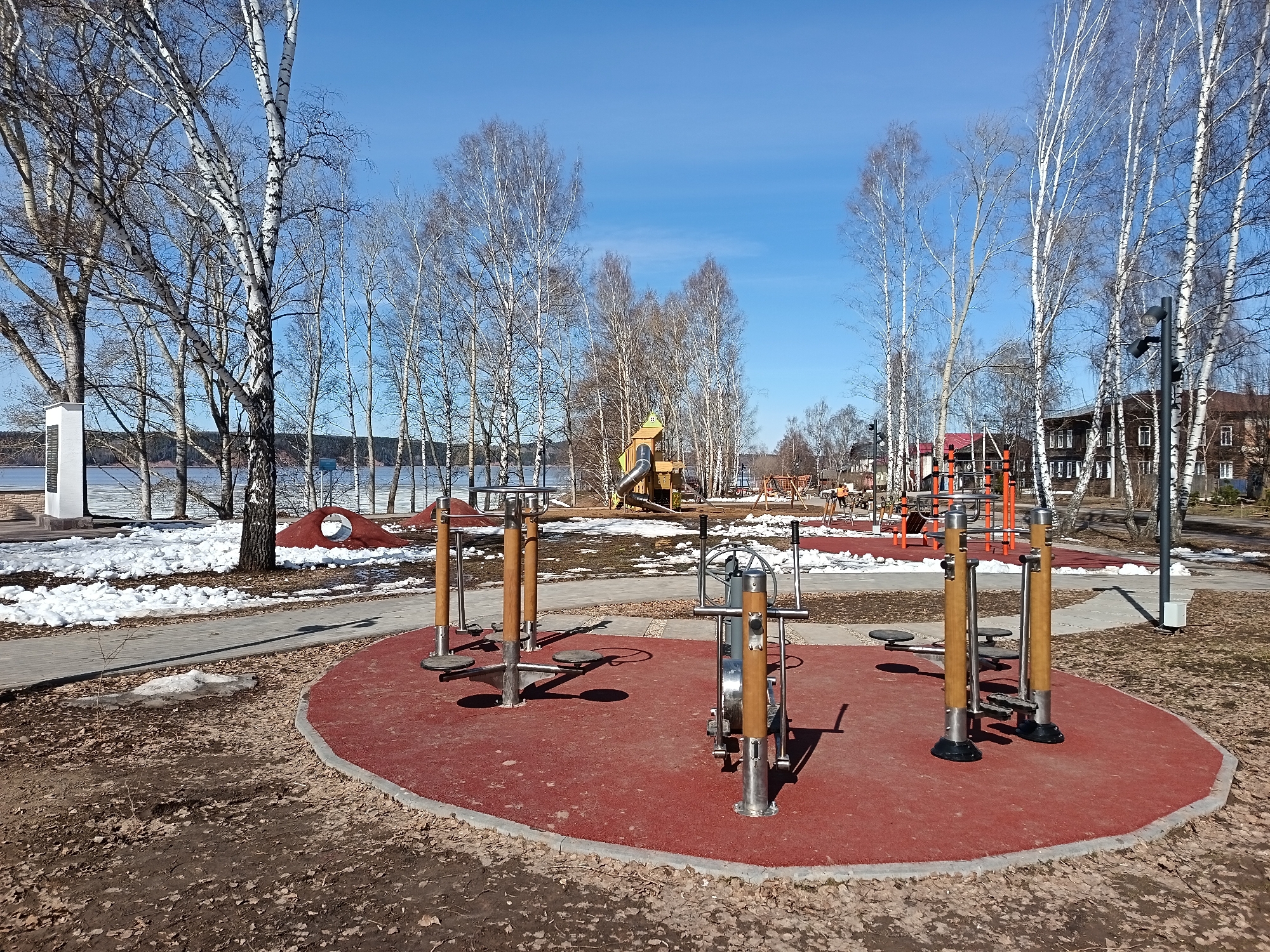 The image shows a public park in Osa, Russia. It is early spring, as evidenced by the melting snow and bare trees. There are outdoor fitness equipment on a red rubberized surface in the foreground. In the background, there's a children's playground with a slide and climbing structures, as well as traditional Russian wooden houses. The park is near a body of water (lake or river). The sky is clear and blue. No people are visible in the scene.