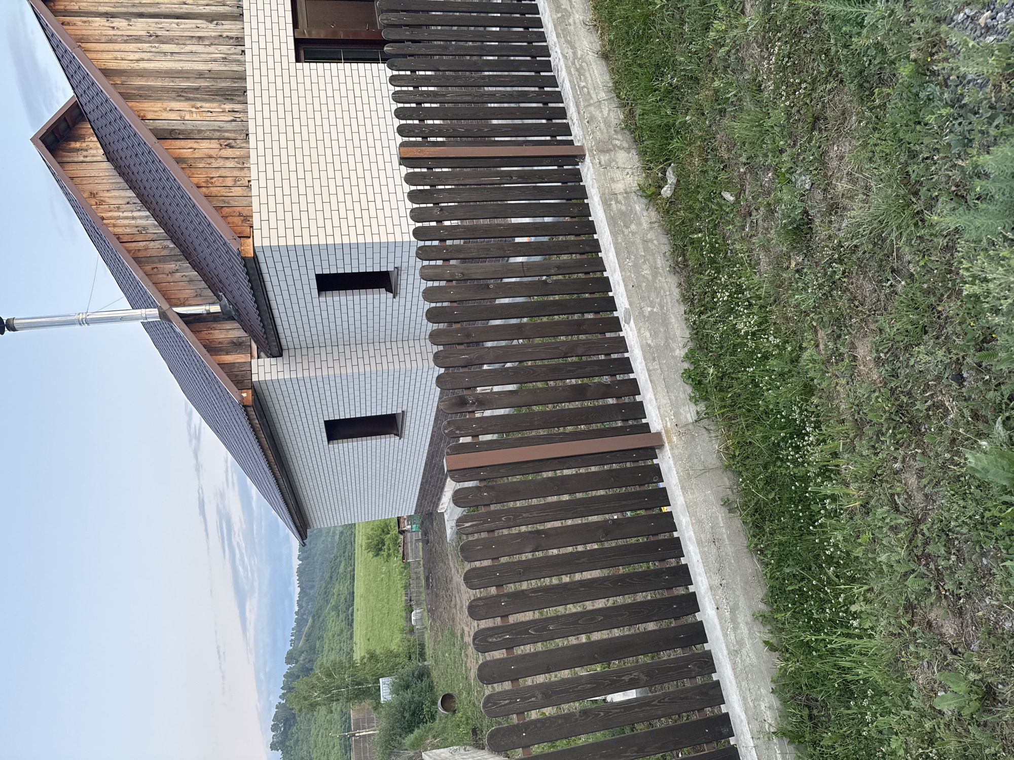 The image depicts the exterior of a house in Soloneshnoye, Russia. The house is constructed with white brick and features a brown shingled roof and wooden gables. A metal chimney extends from the roof. In the foreground, a dark brown wooden picket fence runs along a concrete base. Beyond the fence, a green, hilly landscape is visible under a sky with scattered clouds. The immediate area in front of the fence is covered with grass and weeds.