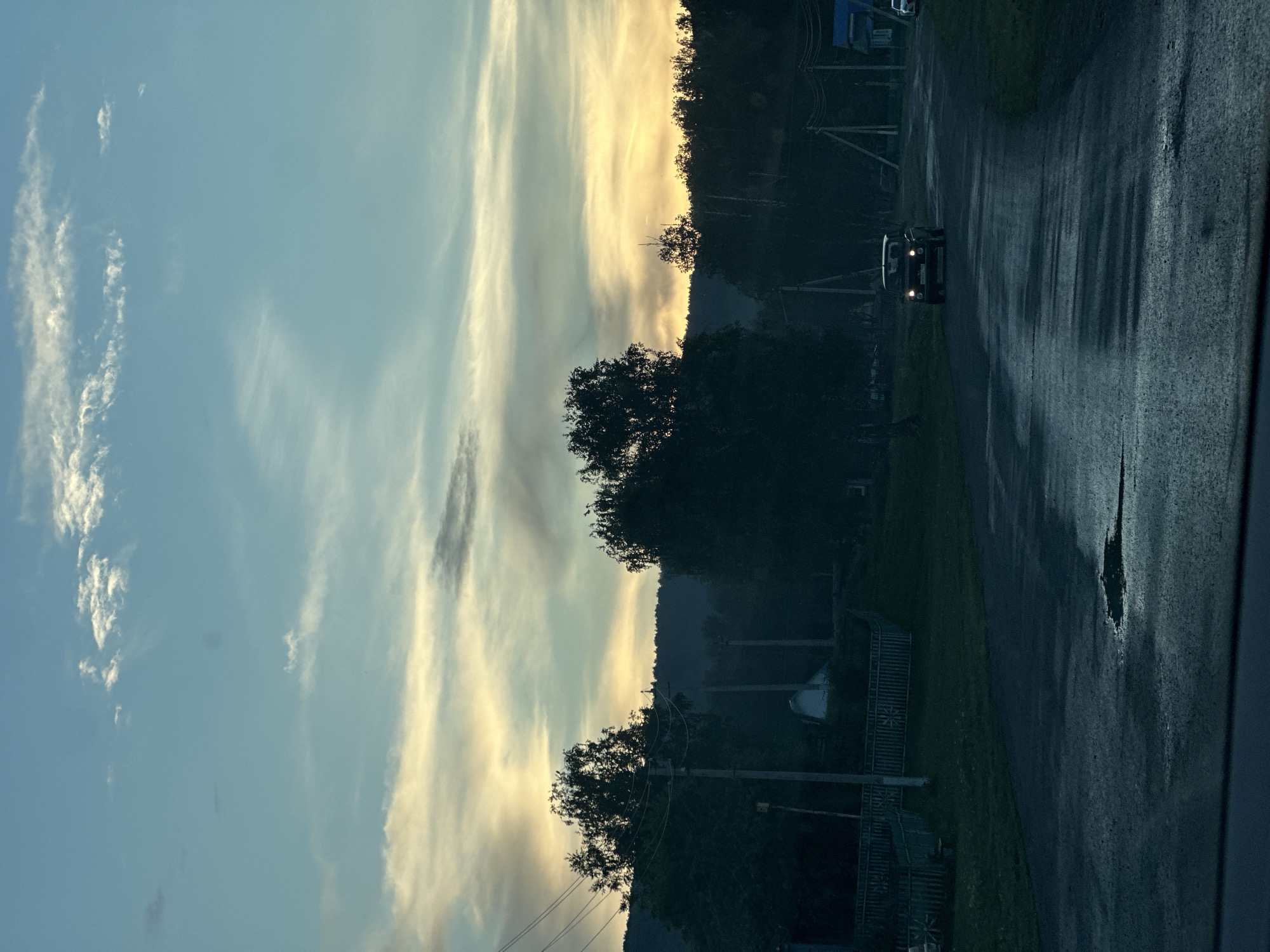 The image depicts a rural or semi-rural road scene at either dusk or dawn, set in the city of Unknown, Unknown. The sky transitions from a light blue with scattered white clouds at the top to a prominent band of yellow and orange hues near the horizon, indicative of a low sun angle, with darker grey clouds interspersed. A line of dark, silhouetted hills or mountains forms the distant background, topped by dense, dark tree lines.
In the midground, several dark green to black trees are visible, obscuring parts of the landscape. Multiple utility poles with power lines crisscross the scene, connecting to unseen structures. On the left side of the road, a light-colored wooden fence with decorative elements is present, behind which a white building with a dark roof is partially visible amidst trees. Patches of green grass are visible adjacent to the road.
The asphalt road in the foreground is dark grey and appears wet, with numerous reflective puddles, including a large, irregular one in the lower center. A dark-colored, older model vehicle, possibly an SUV or off-road car, is positioned in the center of the road, facing the viewer with its headlights illuminated. On the far right, another vehicle is partially visible, parked near a building with a blue roof. The very bottom of the image shows the interior edge of a dashboard or windshield frame. No individuals are visible.