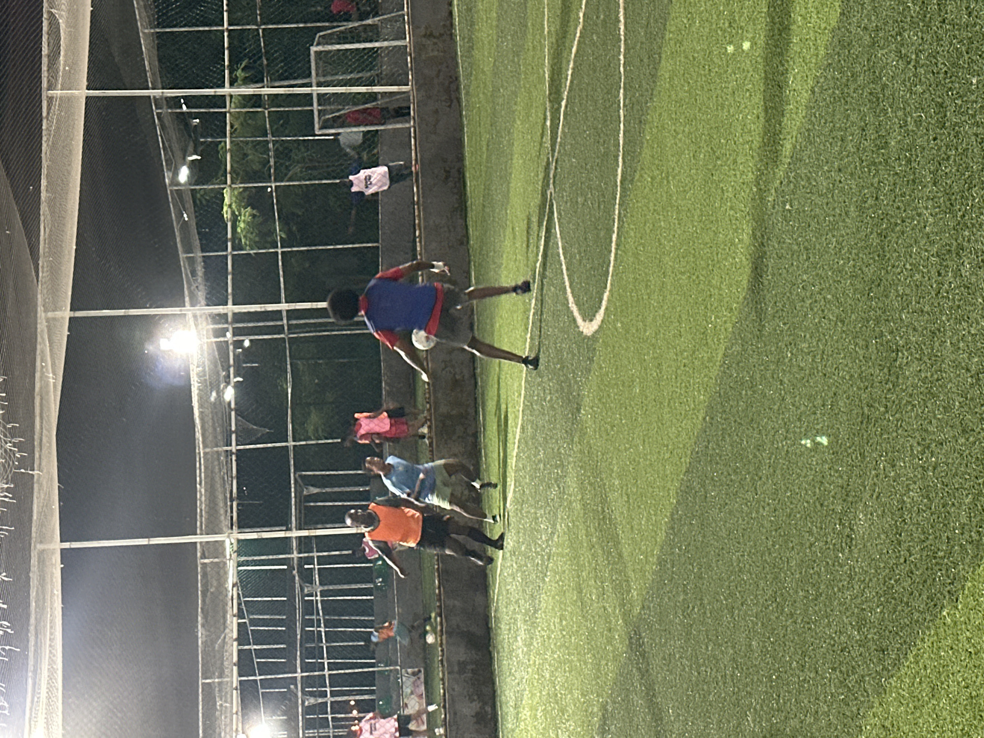The image depicts an outdoor artificial turf football field at night, likely in Abuja, Nigeria, illuminated by artificial lights. Several individuals, presumably playing a match, are visible on the field. In the foreground, a person wearing a blue vest over a red shirt and dark shorts is dribbling a white football. To their left, a person in an orange vest and black shorts is running. Further back, other players in various colored vests can be seen, including pink and blue. The field has white boundary lines and a section of the goal net is visible in the background. The area is enclosed by fencing and netting.