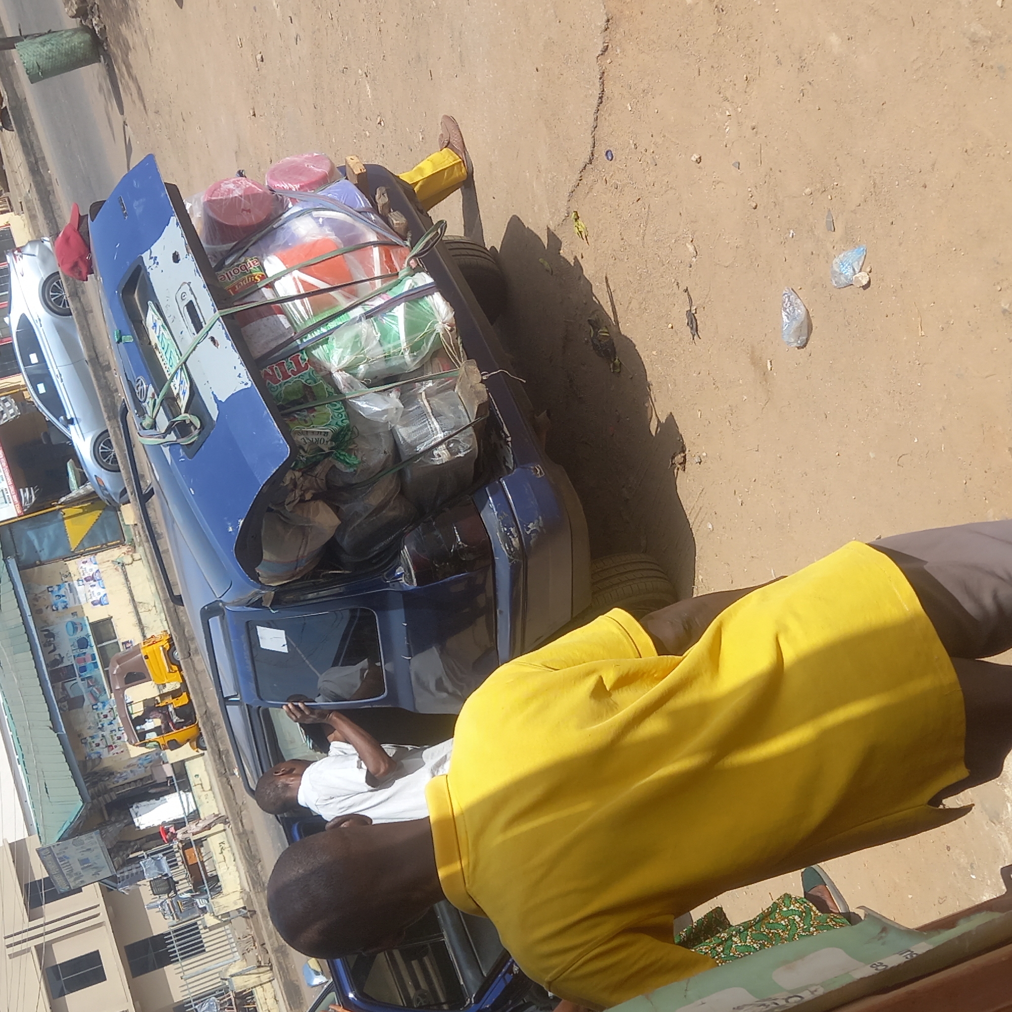This media file captures a street scene, likely in Sardauna, Nigeria, given the context. The image is taken from a slightly elevated perspective, looking down and across a dusty, unpaved road. The bright sunlight suggests it is daytime, casting clear shadows.
In the foreground, the back of a person wearing a vibrant yellow polo shirt and dark pants is visible, facing away from the camera. This person is standing near a heavily loaded blue car.
The blue car, which appears to be a sedan, is the central focus. Its trunk is open, and both the trunk space and the roof are piled high with various goods. On the roof, large red and clear plastic containers are secured with green ropes, along with bundled sacks and other items. Text like "Okpoile" and partially "Super TIM" is visible on some of the cargo. The car's license plate is partially visible, showing some numbers. Another man in a white shirt is visible near the driver's side door of the blue car, seemingly interacting with it or another person inside.
Behind the blue car, a silver or white sedan is parked. Further down the street, on the left side, there are buildings that appear to be shops, some with awnings, and a yellow three-wheeled vehicle (possibly a "Keke Napep") is visible. The ground is largely dirt with scattered small rocks and pieces of trash, including plastic bags. The overall scene depicts a common sight of overloaded vehicles used for transport in some regions.