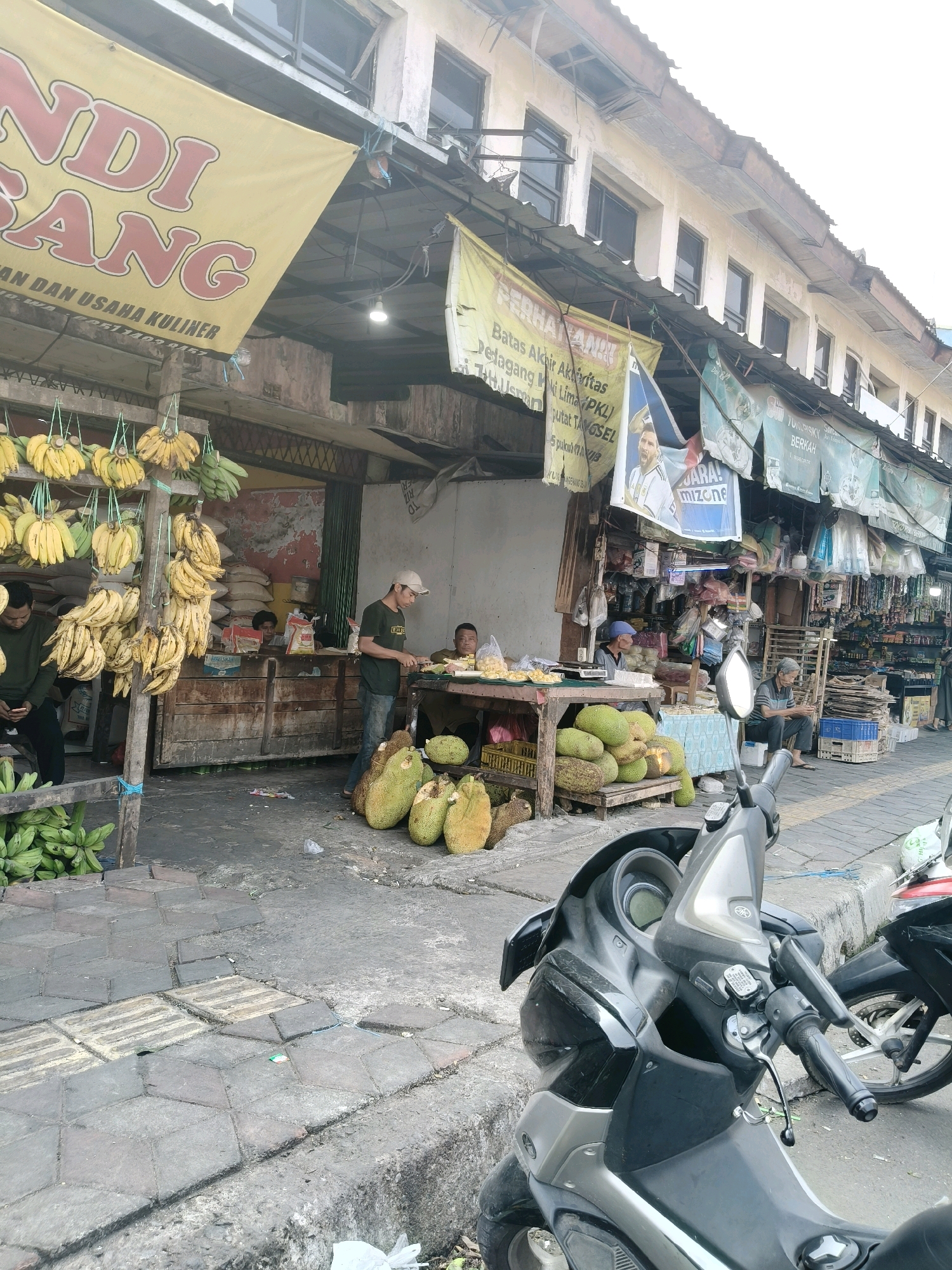 The image depicts a street scene in South Tangerang, Indonesia, likely a market area. In the foreground, a black motorcycle is parked on the sidewalk. The sidewalk is paved with geometric concrete tiles and borders a road. To the left of the motorcycle, a fruit stand displays numerous bunches of yellow bananas hanging from wooden racks and also some green bananas at the bottom. Next to the bananas, there are several large jackfruits piled on wooden crates.
Behind the fruit stand, a wooden stall is visible with people interacting. One person, wearing a dark t-shirt and jeans, is standing and appears to be cutting something on a table. Another person is seated nearby. In the background, there are more market stalls with various goods displayed and other individuals present. Large banners with text and images hang over the stalls, suggesting commercial activity. The buildings lining the street appear to be two stories high with open windows. The overall impression is of a bustling outdoor market.