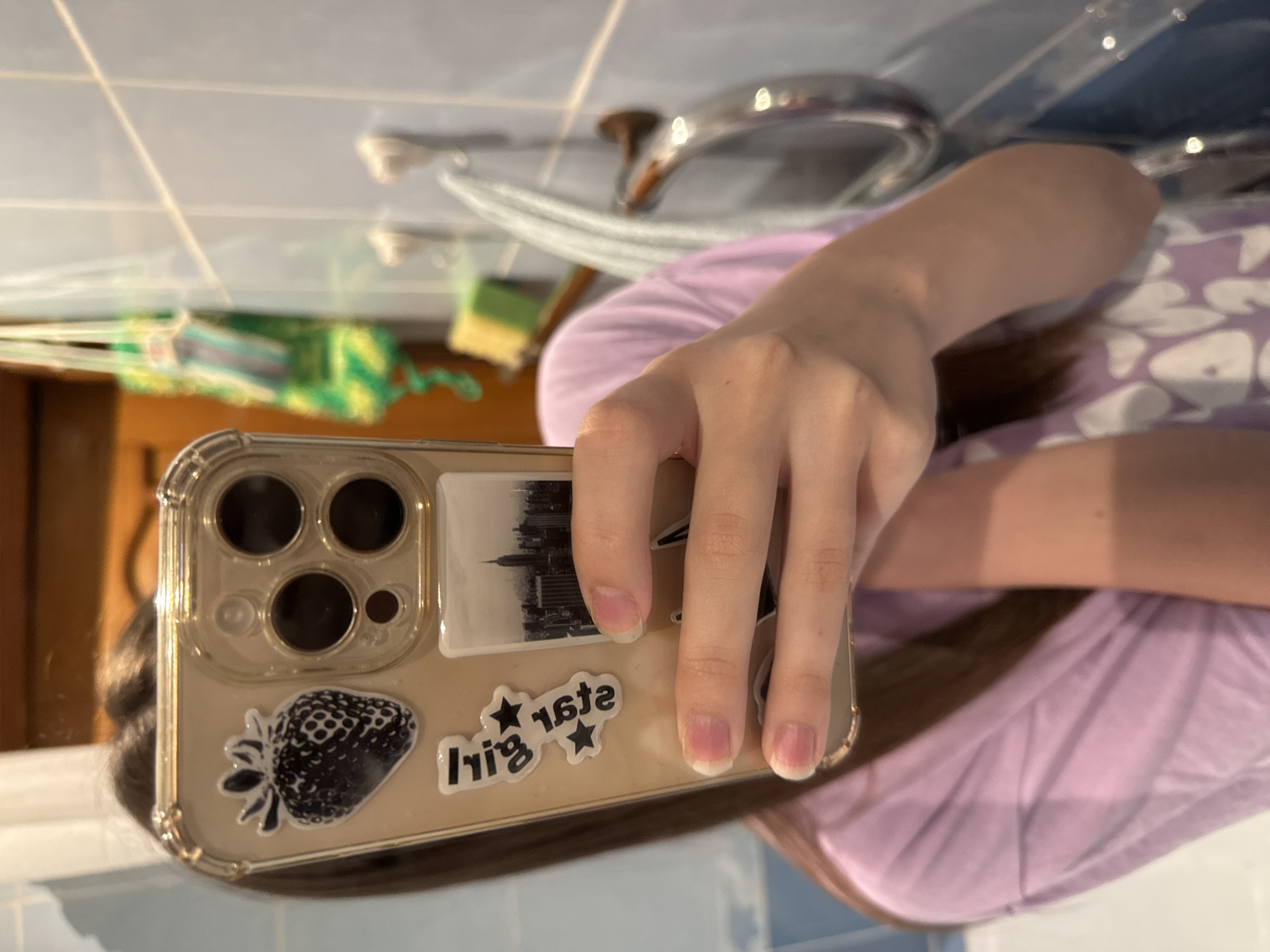 A hand holds an iPhone in front of a mirror in a bathroom. The phone case is decorated with a strawberry sticker, a sticker of a cityscape, and a "star girl" sticker. The person holding the phone is wearing a purple patterned shirt. The bathroom features blue and white tiles, a sink with a chrome faucet, and a green cleaning item in the mirror's reflection. The city of Unknown, Unknown is not discernible from this image.