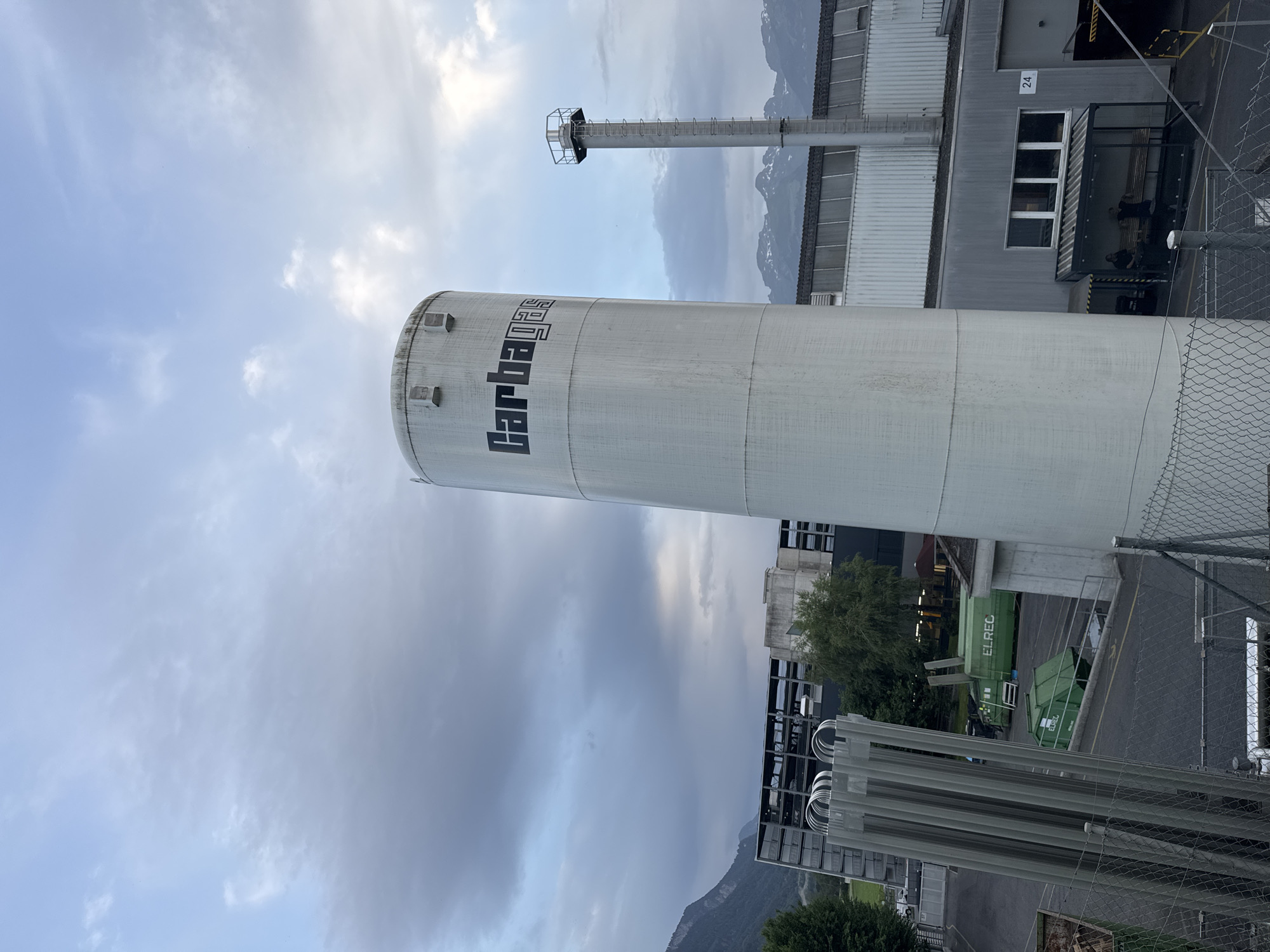 The image shows an outdoor industrial scene, likely in Nendeln, Liechtenstein, with a large, white, cylindrical storage tank prominently featured in the foreground. The tank has the word "Carbagas" printed in large, black, stylized letters on its upper section. To the right of the main tank, a tall, slender industrial chimney with a ladder and a platform at the top is visible. In the background, a multi-story industrial building with a metal facade and multiple windows stretches across the frame. Mountains can be seen in the distance, suggesting a location with a mountainous terrain. Several green waste bins, one with "ELREC" printed on it, are scattered in the paved area. A fence runs along the bottom of the image. The sky is partly cloudy, with a mix of light and dark clouds, indicating overcast or partly sunny conditions. The lighting suggests it is either early morning or late afternoon.