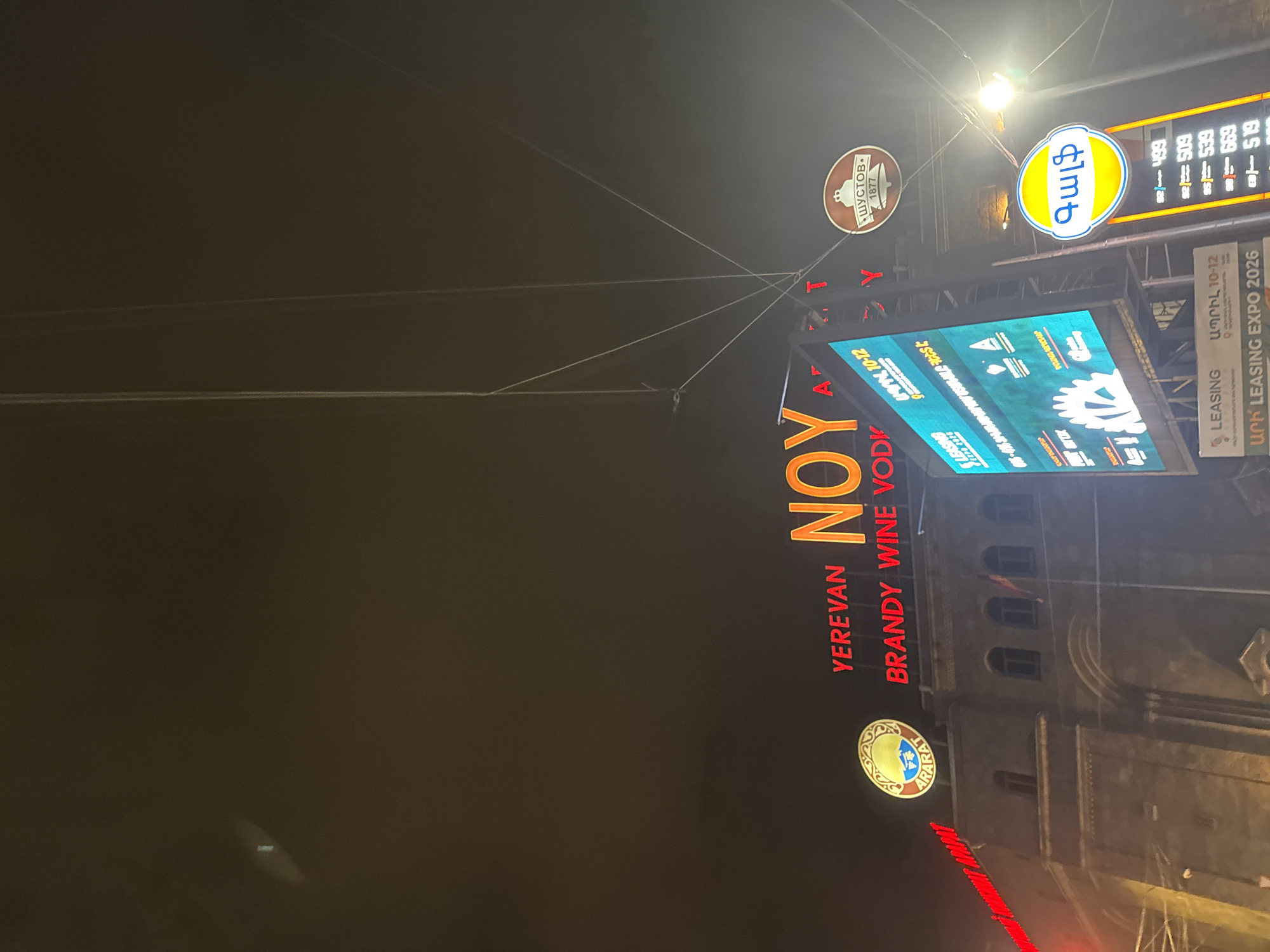 The image shows a nighttime street scene in Yerevan, Armenia, dominated by several illuminated advertising signs. Prominently featured is a large red sign advertising "Yerevan Noy Brandy Wine Vodka." Above this, there's a digital display screen showing commercial content. Additional signs above include a circular sign with the word "Gulf" in Armenian script, and another for "Shustov 1877." The architecture suggests a historic building in the background. There is also an advertisement about leasing expo 2026.
