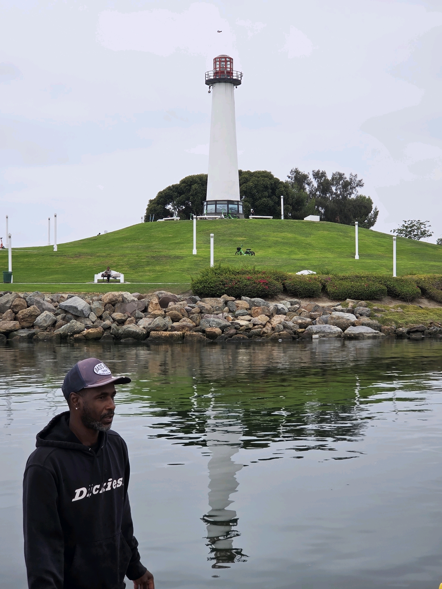 This image captures a scenic view of the Long Beach Peninsula in Long Beach, California, United States of America. In the foreground, a man wearing a black hoodie with "Dickies" printed on it and a dark baseball cap stands facing to the right, with his reflection visible in the calm water. The water itself occupies the lower portion of the frame and reflects the surrounding environment.
Behind the man and the water, a rocky embankment separates the water from a lush green hill. Several large boulders and stones form the embankment. Atop the hill, a tall, white lighthouse with a red top stands prominently against a cloudy sky. The lighthouse is a focal point on the grassy slope, which is dotted with trees and some white poles. In the middle ground, a person is seated on a white bench on the hill. Two green electric scooters are also visible on the grass, near the base of the lighthouse. The overall atmosphere is serene and coastal.