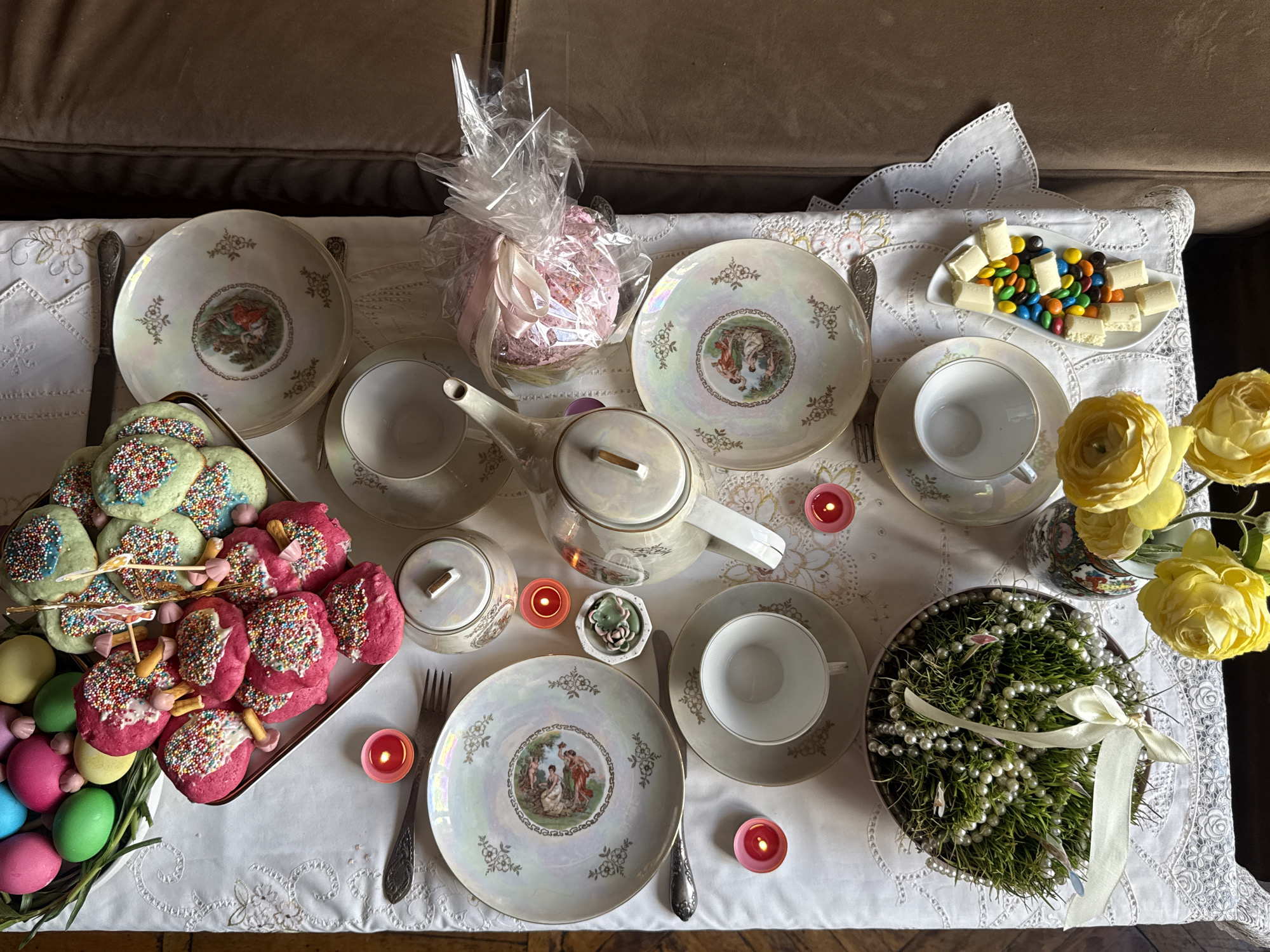 A high-angle shot shows a table set for tea, likely indoors, although there are no visual clues to pinpoint the exact location in Yerevan, Armenia. The setting includes four place settings with antique porcelain plates, teacups, and silverware. A white embroidered tablecloth covers the table.
A silver tray holds stacks of cookies with colorful sprinkles and decorated eggs, suggesting an Easter or springtime theme. In the center of the table, a teapot and sugar bowl accompany a small dish with a succulent plant. There are also small pink candles distributed around the table. A dish of white chocolate and M&Ms sits next to a vase of yellow flowers. Another decorated cake is wrapped in cellophane with a pink ribbon.
To the right of the frame is a decorative pot containing green grass adorned with white pearls and a ribbon. The edge of a brown couch is visible along the top of the frame.