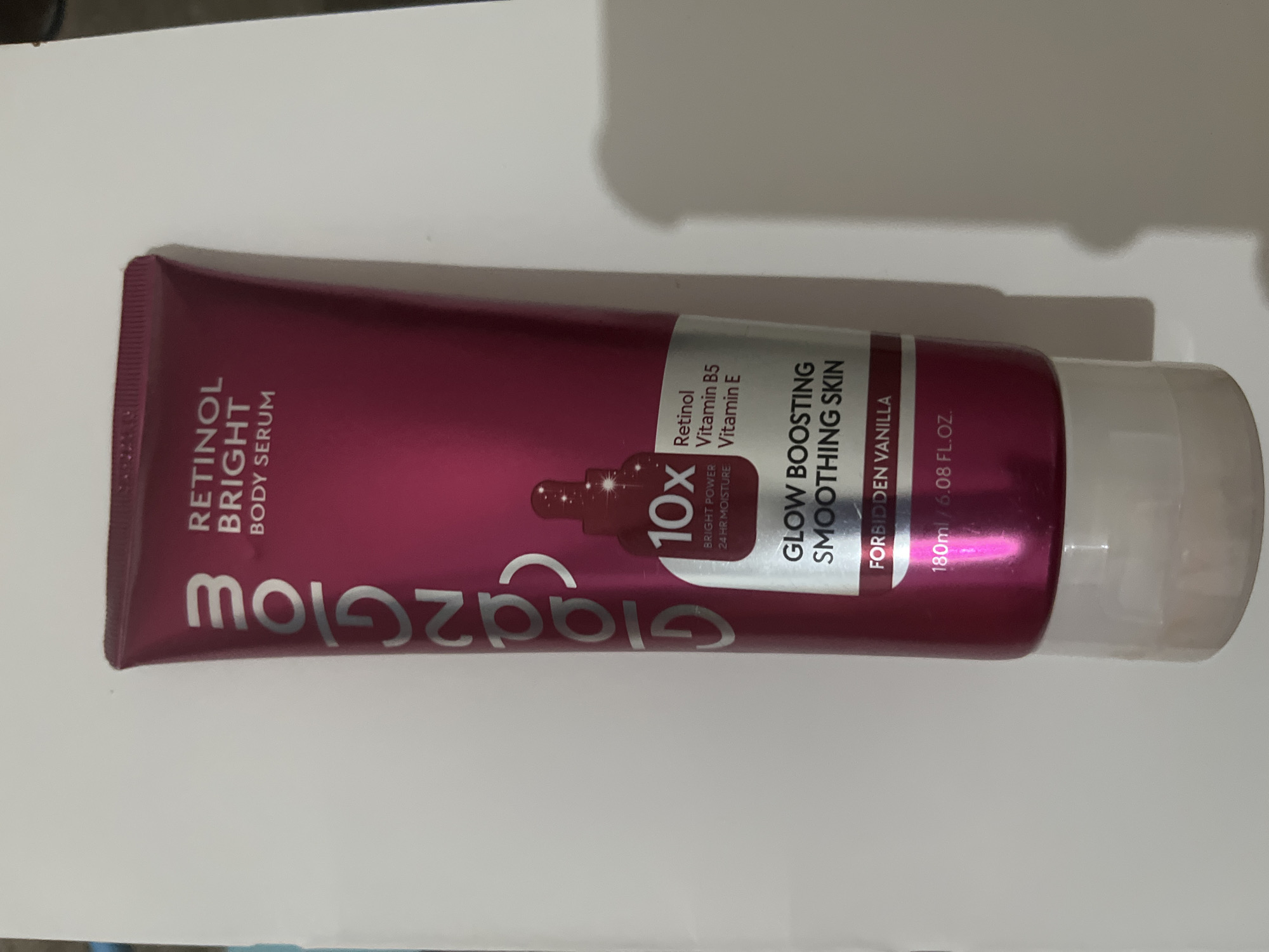 A full shot shows a tube of Glazeglow Retinol Bright Body Serum standing upright against a white background. The tube is predominantly magenta with silver lettering. The Glazeglow logo is vertically oriented on the left side of the tube. Text on the right side of the tube reads "RETINOL BRIGHT BODY SERUM" at the top, followed by a graphic of a dropper bottle with "10X" superimposed on it. Below the dropper graphic, it says "BRIGHT POWER 24 HR MOISTURE," and "Retinol Vitamin B5 Vitamin E." Further down, the tube advertises "GLOW BOOSTING SMOOTHING SKIN" and "FORBIDDEN VANILLA." At the bottom, the volume is listed as "180ml / 6.08 FL.OZ." The product appears to be new and undamaged. The lighting is even, with a subtle shadow cast to the right of the tube. The image is likely a product photo, and the location within Meliau Hulu, Indonesia is not discernible from the visual information provided.