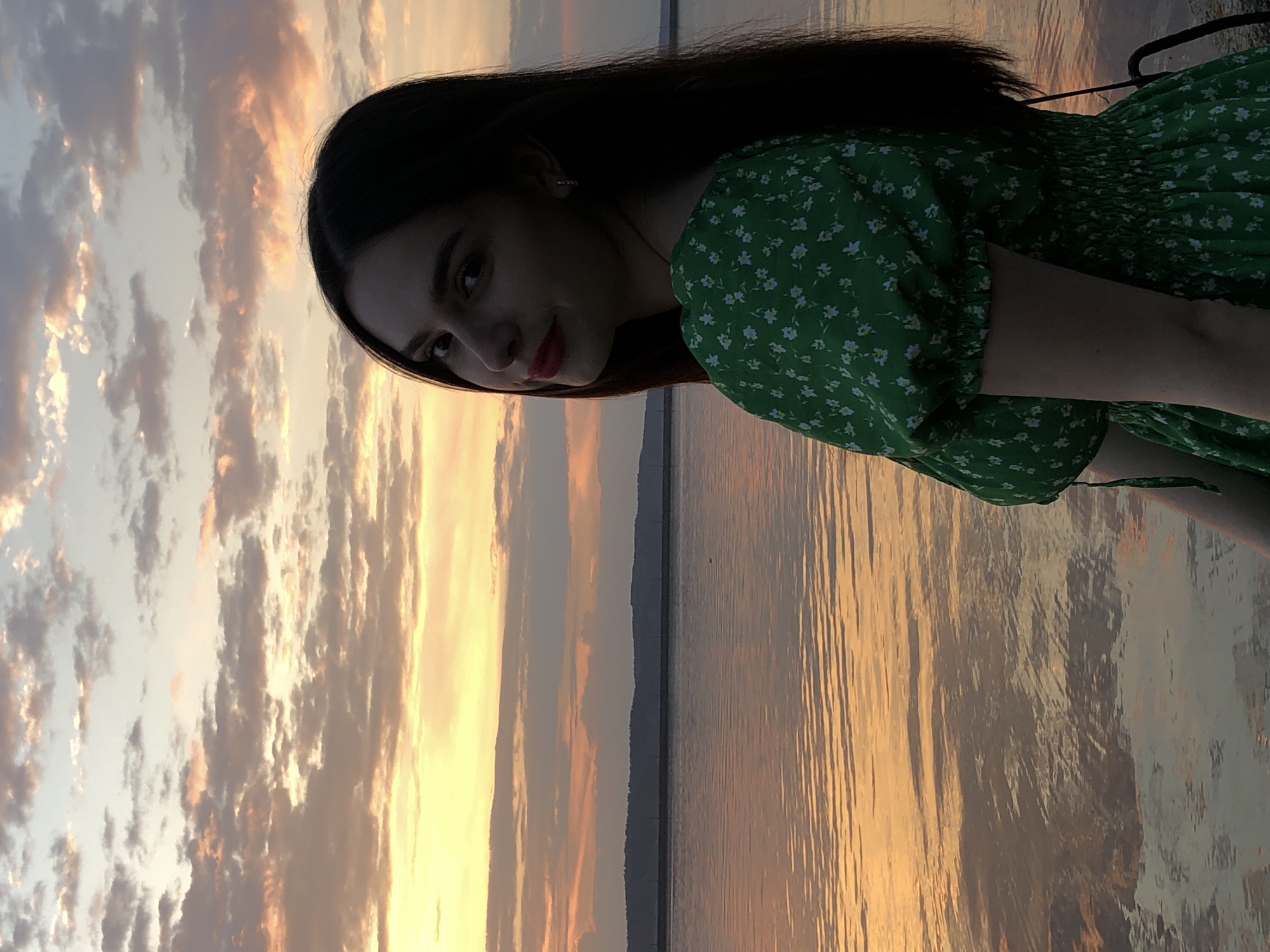 A young woman with dark hair and a green floral dress stands against a backdrop of a sunset reflected in a body of water. The sky is filled with clouds tinged with orange and yellow hues. The woman appears to be standing by a lake or large river, reflecting the sunset. The scene's location is Chaikovsky, Russia.