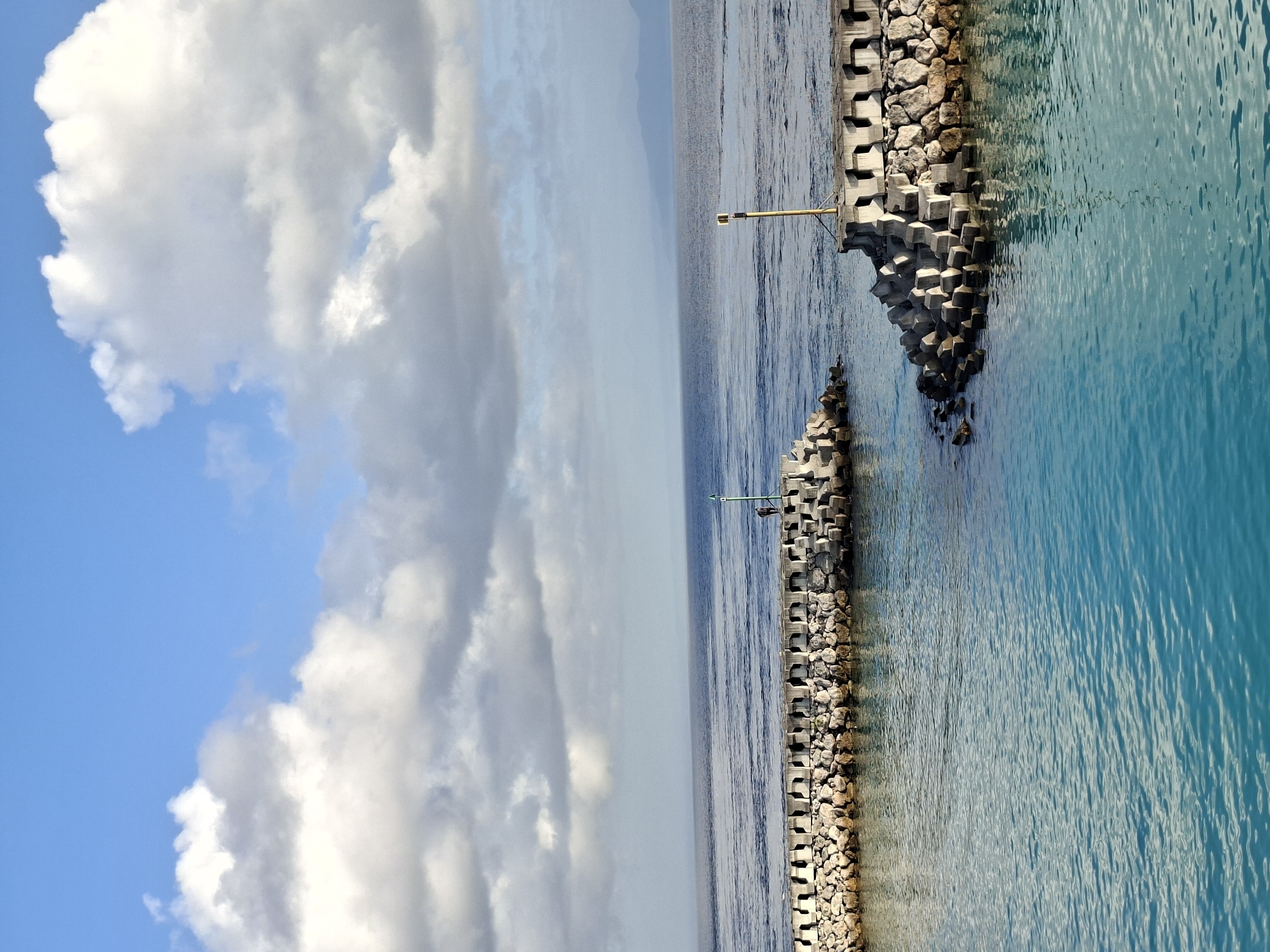 The image depicts a body of turquoise water in front of a breakwater constructed from concrete tetrapods and large stones. A single person, dressed in dark clothing, stands on the breakwater on the right side of the image, near a yellow pole with a navigational marker. In the distance, the sea stretches to a hazy horizon where landmasses are faintly visible. The sky above is a mix of blue and white clouds. The scene appears to be located in Nusa Penida, Indonesia, likely a coastal area with a harbor or marina.