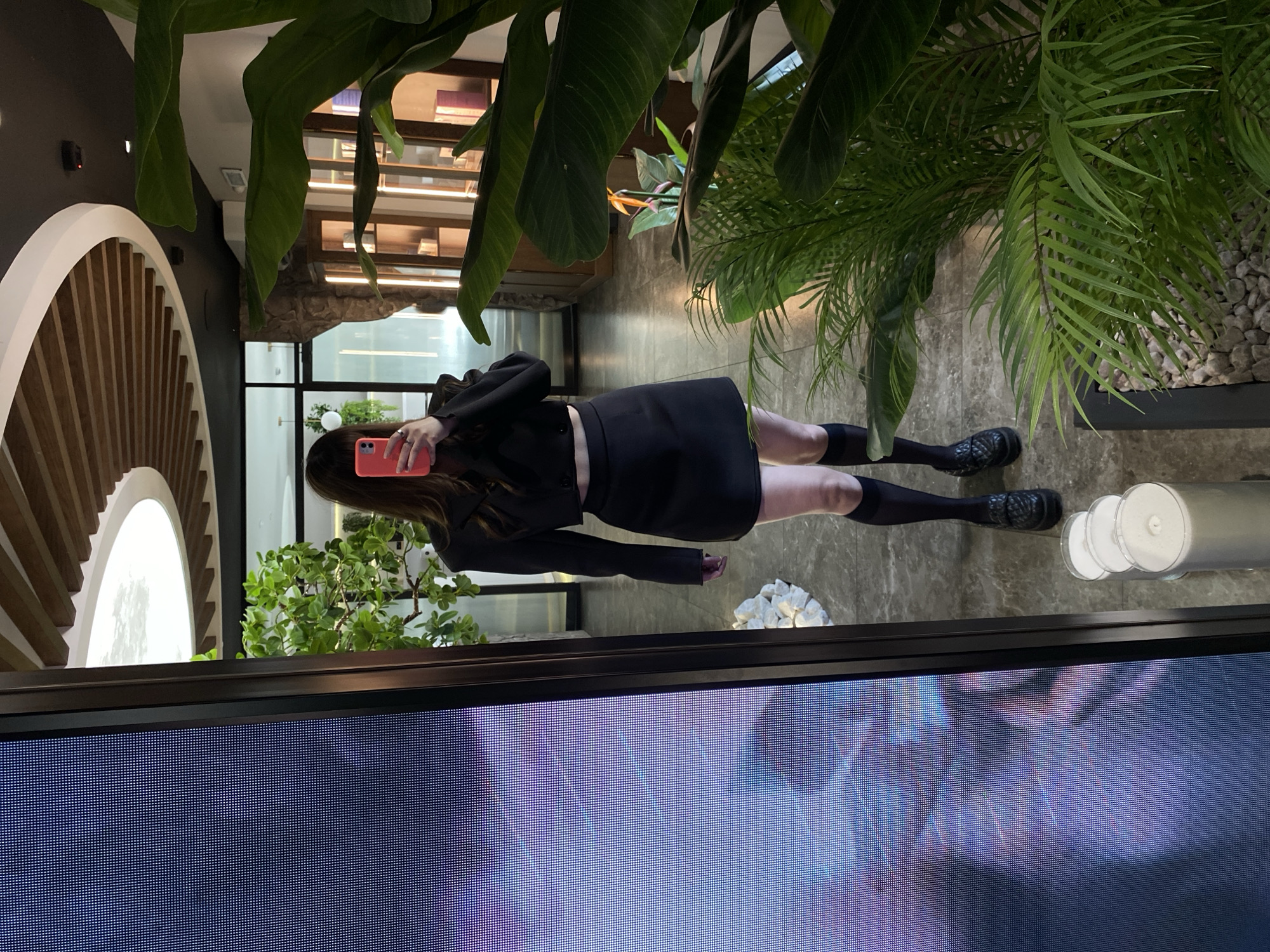 A woman is holding a pink phone, standing indoors, apparently in front of a screen displaying a colorful abstract pattern. She is wearing a black blazer, skirt, socks, and shoes. The surroundings include lush greenery and architectural elements that feature a partially visible wooden-slatted, semi-circular structure. The reflection on the screen hints at a location, potentially the city of Votkinsk, Russia.
