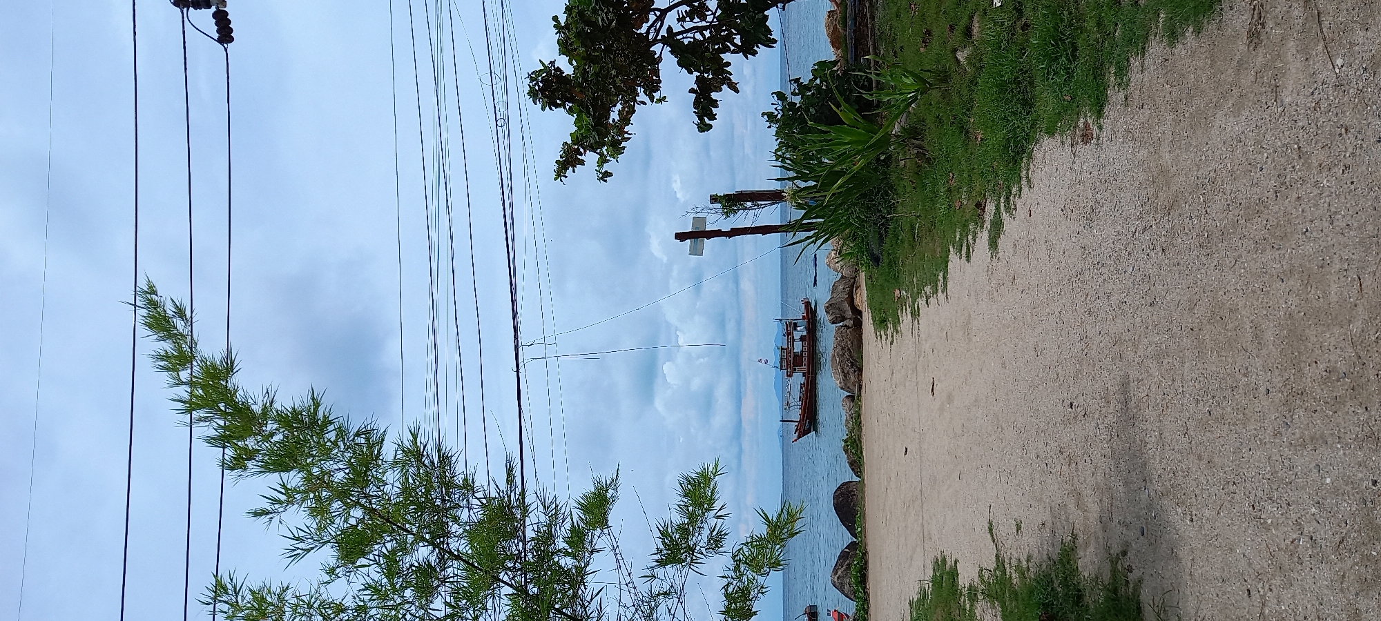 The image shows a coastal scene in Baan Chaloklum, Thailand. In the background, a body of water, presumably the sea, is visible with fishing boats anchored near the shore. The sky is overcast with clouds. A sandy beach or pathway runs along the right side of the frame, transitioning into a paved or gravelly area in the foreground. Along the shoreline, there are rocks and green vegetation. On the left side of the frame, there are trees and power lines. A wooden pole with signage is also visible near the water.