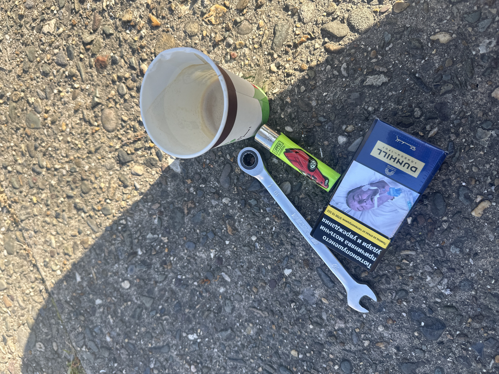 The image shows a littered scene on a concrete ground with embedded pebbles. A disposable coffee cup with coffee inside lies on its side. Next to it are a green Clipper lighter with a red car on it, a silver-colored wrench, and a pack of Dunhill cigarettes with a picture of a sick person on the pack, possibly as a warning about the dangers of smoking. The wrench bears the markings "10" and "ARKSIDE". The cigarette pack also contains text in a language other than English, potentially Bulgarian. The scene is partially in sunlight, casting a shadow on the left side of the image. The location is unknown and nothing suggests it is in Nivelles, Belgium.