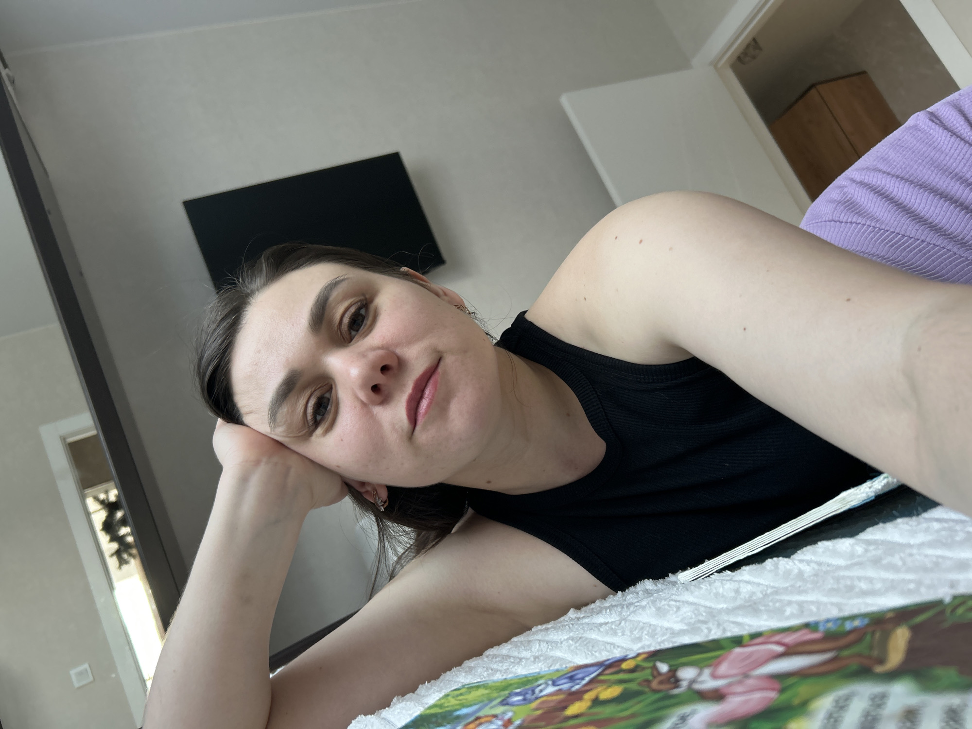 A young woman with dark hair is lying on a bed, resting her head on her left hand. She is wearing a black tank top. A book lies open in front of her. In the background is a television mounted on the wall. A door and what appears to be a cabinet are visible. The photo was taken in what looks like an apartment room. There is no information about the location of the city of Chaikovsky in Russia.