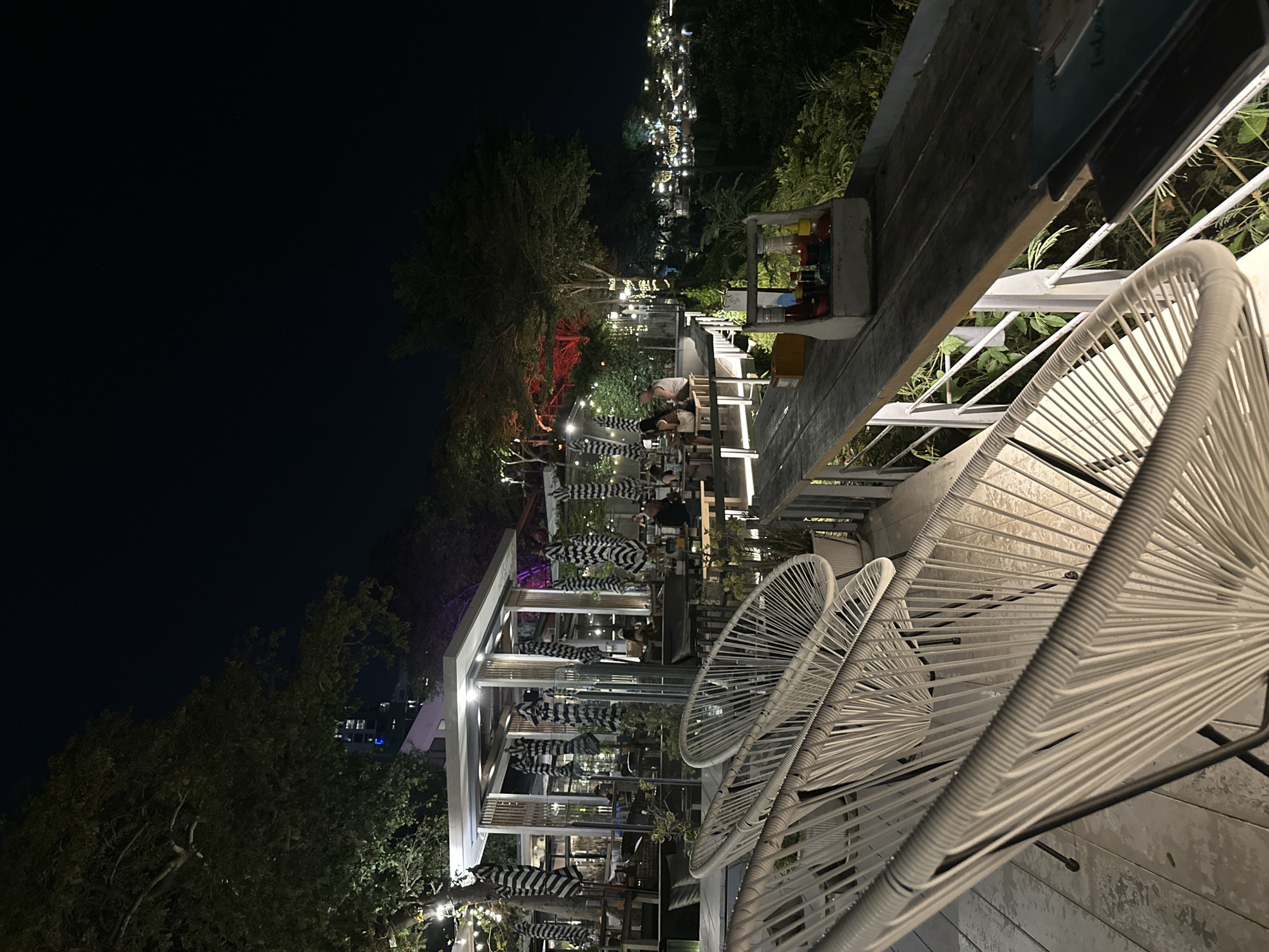 The image depicts an outdoor bar or restaurant scene at night in Pattaya City, Thailand. The view is taken from a balcony or elevated seating area, with white wicker chairs in the foreground occupying the right side of the image. The background shows a restaurant with a multi-level outdoor seating area, illuminated with lights. Diners are visible at various tables, and striped umbrellas are present to provide shade. The overall ambiance is relaxed and social. To the left, there is a tree and a building in the distance, with the night sky filling much of the upper portion of the image.