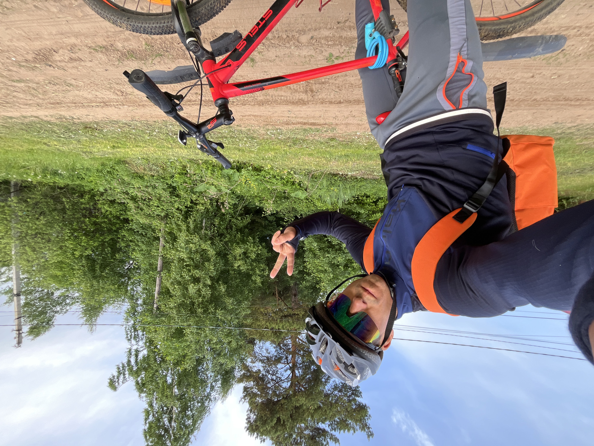 A man in a helmet and visor is posing with a peace sign next to a red and black mountain bike in Chaikovsky, Russia. The bike has a blue lock. The man wears grey and blue athletic clothing with orange accents and an orange bag. The background shows a dirt road, green foliage, trees, power lines and a blue sky.