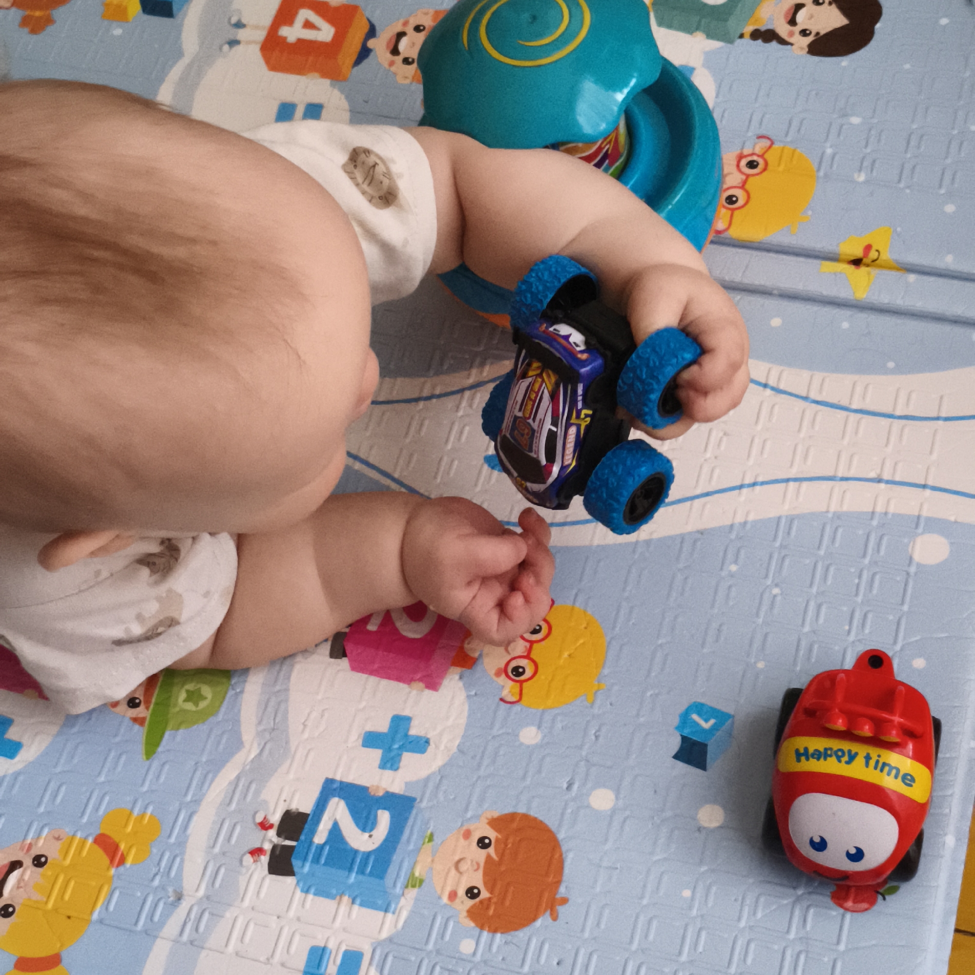 A baby lies on a padded play mat with colorful designs in a room in Chaikovsky, Russia. The baby wears a white diaper with animal prints and is surrounded by several toys, including a blue and black toy car with large blue tires, a red toy car labeled "Happy time", and a blue and green spinning toy. The play mat features cartoon characters and educational elements like numbers and addition signs. The scene is viewed from an elevated perspective, focusing on the baby and the surrounding play area.