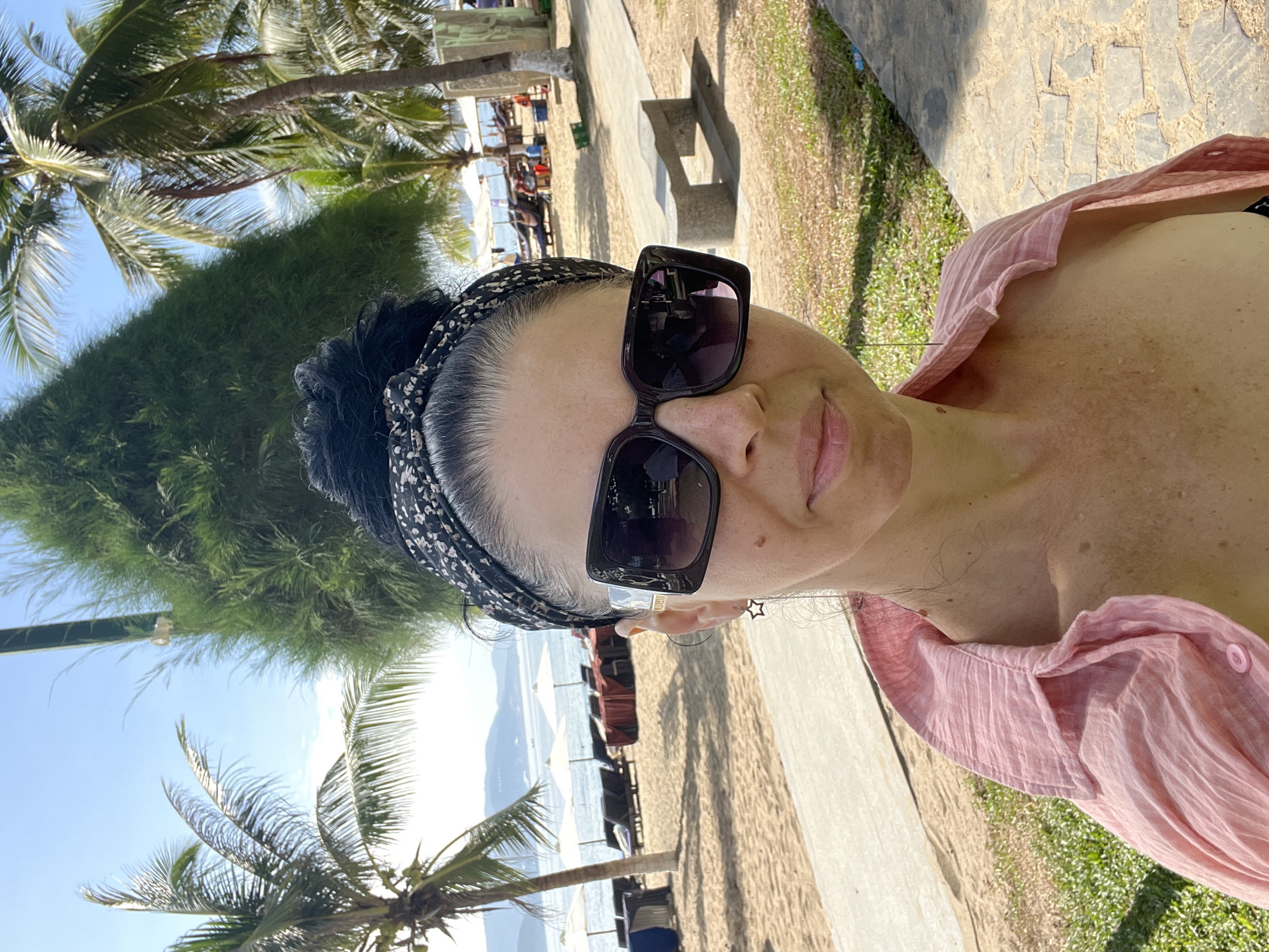 A woman with dark hair styled in a bun and a black and white bandana stands on a beach in Nha Trang, Vietnam. She is wearing large, square, dark sunglasses and a light pink, button-up shirt. The beach is lined with palm trees and lounge chairs. In the background, the ocean meets a hazy mountain range. The sun is shining, casting shadows on the sandy ground.