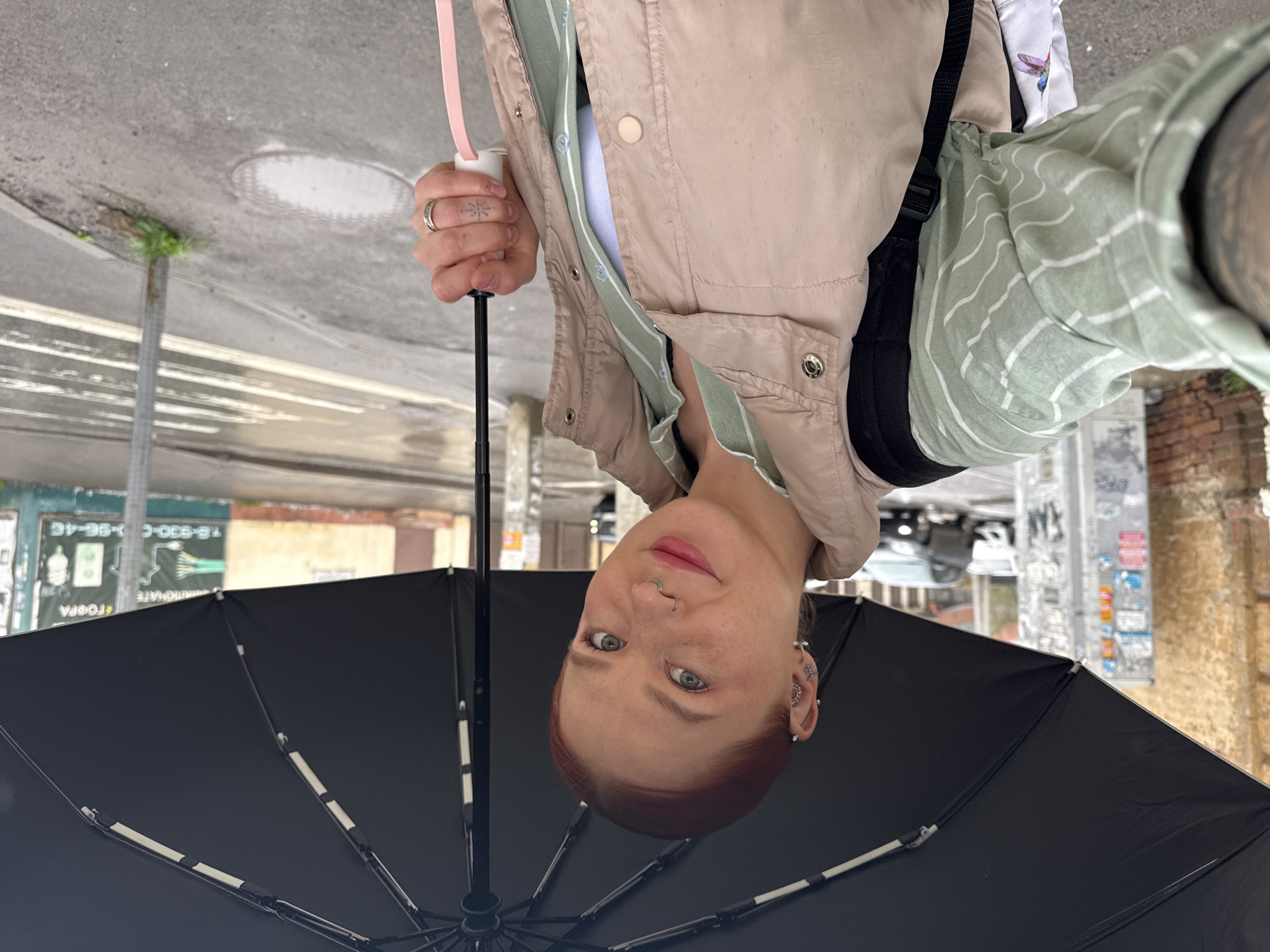 A woman holds a closed black umbrella while standing outside. The location appears to be Rostov-on-Don, Russia, as inferred from a sign in Russian visible in the background. The woman has red hair, light skin, multiple piercings in her ear and nose, and wears a light-colored jacket over a striped shirt. She also has a tattoo on her hand. The background shows an urban street setting with buildings, a street light, and other typical city elements.