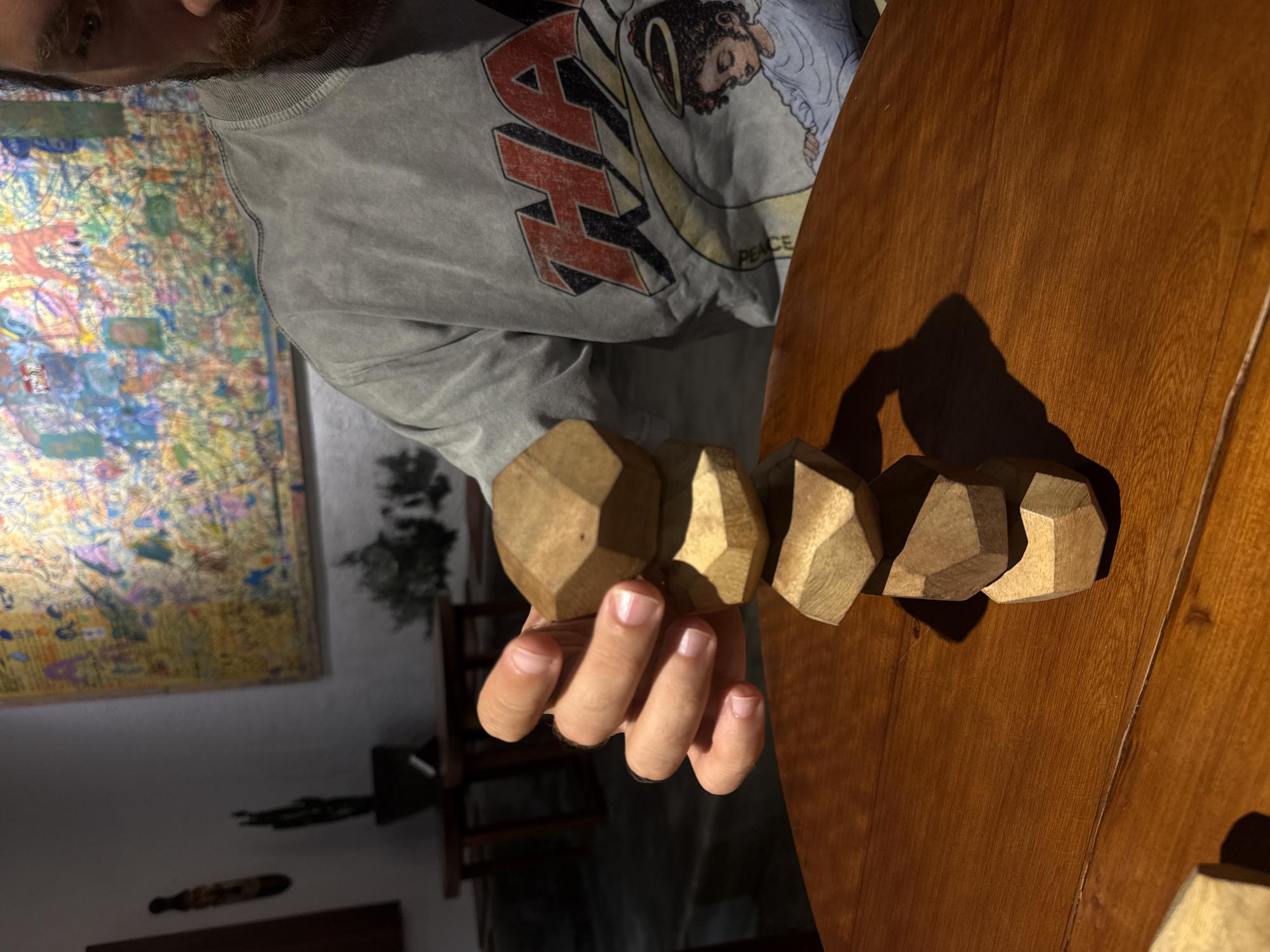The image shows a person's hand holding several wooden geometric blocks over a wooden table. The person is wearing a grey sweatshirt with a graphic print featuring a drawing of a person with wings and the word "PEACE". In the background, a colorful abstract painting hangs on a white wall. The setting appears to be indoors, possibly a home or studio, with natural light casting shadows. The time of day is difficult to determine, but the lighting suggests it might be late afternoon or early evening. There are no clear indicators of weather. The overall mood is calm and contemplative, with the focus on the tactile quality of the wooden blocks and the artistic elements in the background. The location context of Da Lat, Vietnam, is provided but not visually evident in the image itself.