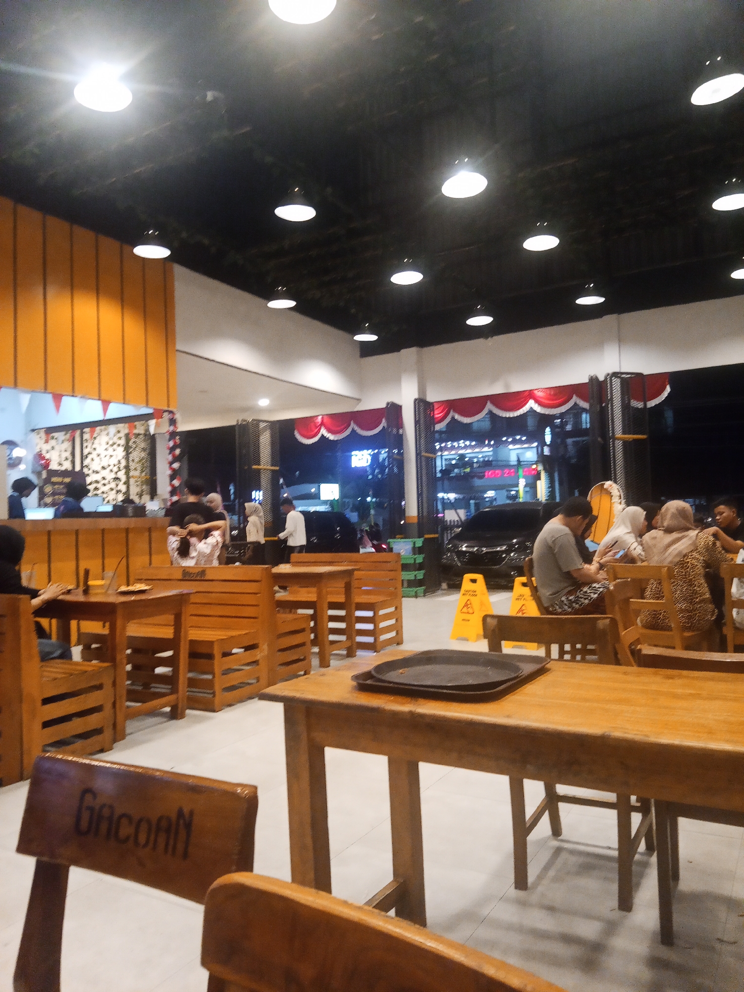 The image captures the interior of a restaurant in Lubuklinggau, Indonesia, during nighttime. The environment is dimly lit with artificial lighting emanating from numerous overhead fixtures. Wooden tables and chairs are arranged throughout the dining area, some occupied by patrons. In the foreground, a wooden chair with "Gacoan" inscribed on its back is visible. In the background, a counter area with staff can be seen, and beyond that, a street scene with cars and buildings is visible through an open doorway. The overall atmosphere appears casual and busy.