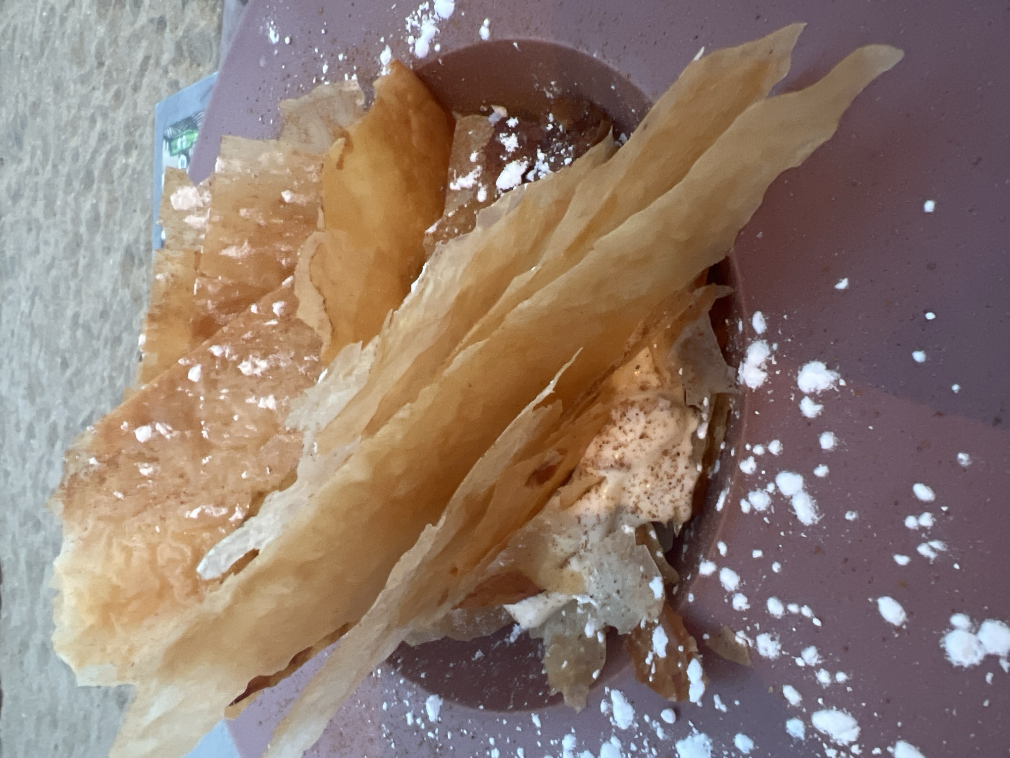 A close-up shot shows a dessert on a purple plate. The dessert is topped with thin, crispy, golden-brown layers of pastry, possibly phyllo dough, dusted with powdered sugar. Beneath the pastry, there appears to be a creamy filling, also sprinkled with cinnamon and powdered sugar. The plate is situated on a sandy surface, suggesting an outdoor setting, possibly a beachside restaurant in Eretria, Greece, given the location context. In the background, out of focus, there is a suggestion of a table with a menu or placemat.