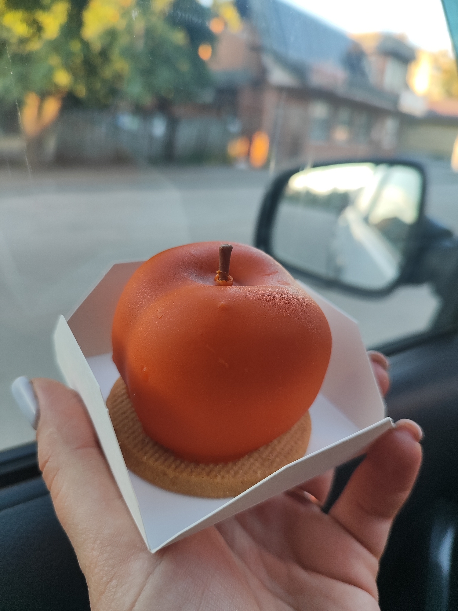 A person's hand holds a small, orange, apple-shaped dessert in a white paper container. The dessert is placed on a round cookie. The dessert has a small brown stem. The background is out of focus and shows trees, buildings, and a car's side mirror. The scene appears to be inside a car during the day.
