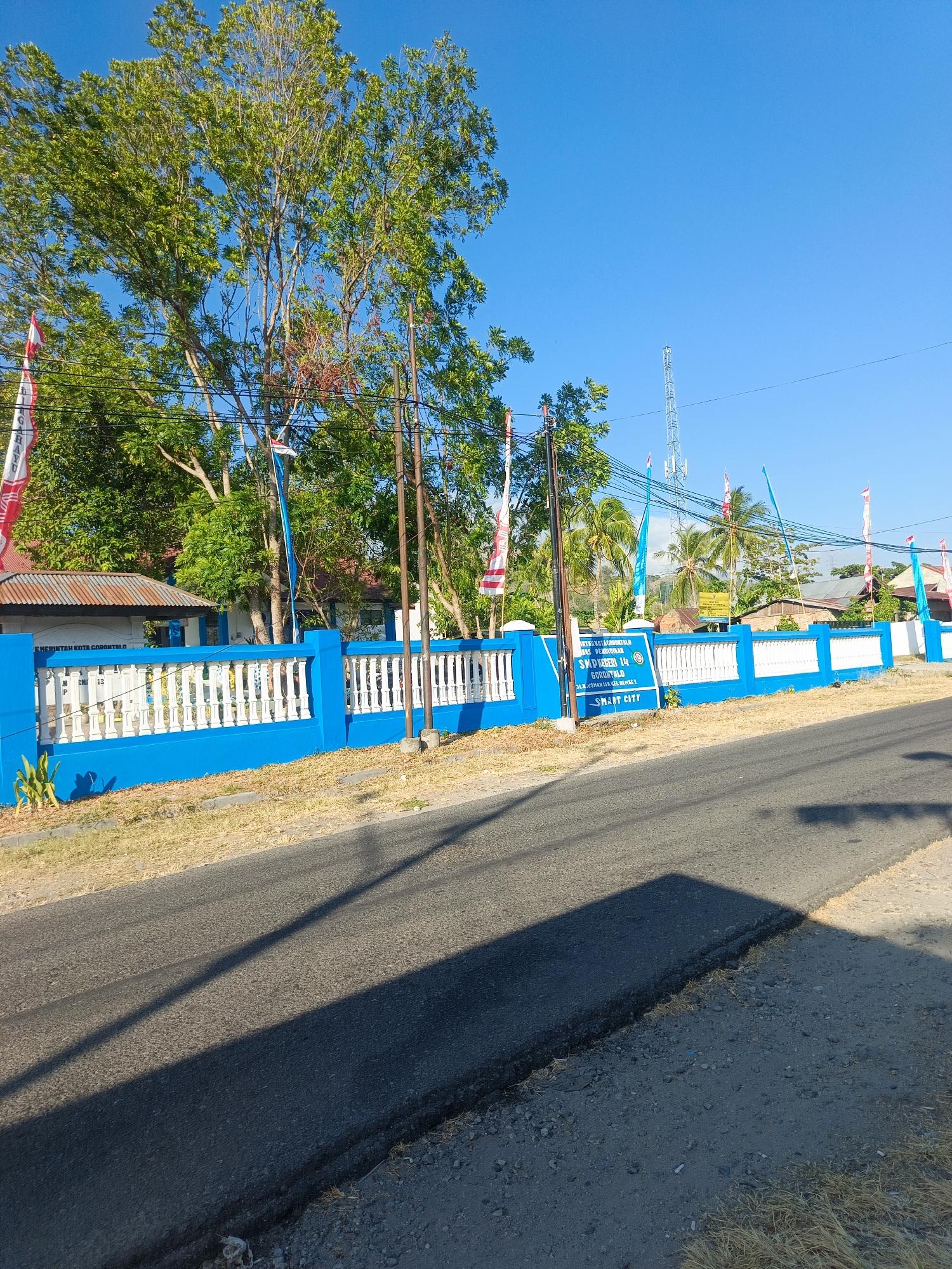 The image depicts a street scene in Gorontalo, Indonesia, with a bright blue sky overhead. In the foreground, a paved road runs diagonally across the frame. The road appears to be in moderate condition with some rough edges and debris on the shoulder. A prominent shadow from an unseen object stretches across the road.
Behind the road is a low, dry grassy area and then a bright blue wall topped with white balusters, forming a fence. Several Indonesian flags, red and white, are visible, some attached to poles along the fence and others flying freely against the sky. Behind the fence, there are trees with green foliage, and some buildings with corrugated metal roofs. A white sign with blue lettering is mounted on the fence, indicating "SMP NEGERI 14 GORONTALO" and the phrase "SMART CITY". Further in the background, a tall telecommunications tower and more buildings and trees are visible. The overall impression is a sunny day in a semi-urban or suburban area.