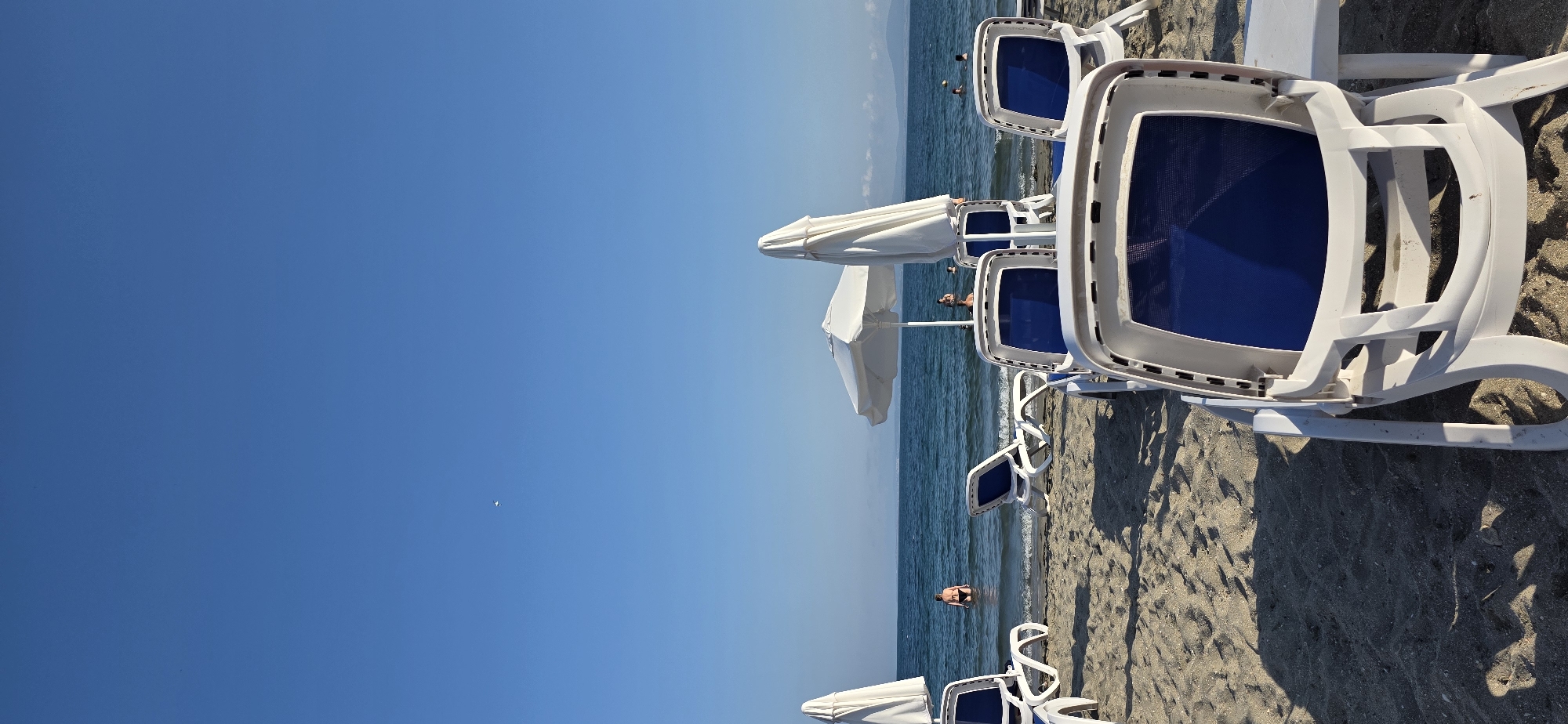 This is a vertical, eye-level, outdoor, daytime photograph taken on a sandy beach in Pomorie, Bulgaria. The image captures a serene beach scene with the clear blue sea in the background. In the foreground, several white plastic lounge chairs with blue fabric seats are arranged on the sand, some facing the sea and others facing away from the viewer. One chair in the immediate foreground is prominently in focus, showing its detailed texture and construction.
To the left and center of the image, large white beach umbrellas are visible, one fully open and the other closed and standing upright. In the middle ground, the calm sea stretches to the horizon, with a few people wading or swimming in the water. One person in a dark swimsuit is standing in the shallow water near the shore.
The sky above is a vast expanse of uninterrupted, bright blue. The sand is light brown with visible footprints and textures. The overall impression is one of a peaceful day at the beach, with ample space for relaxation.